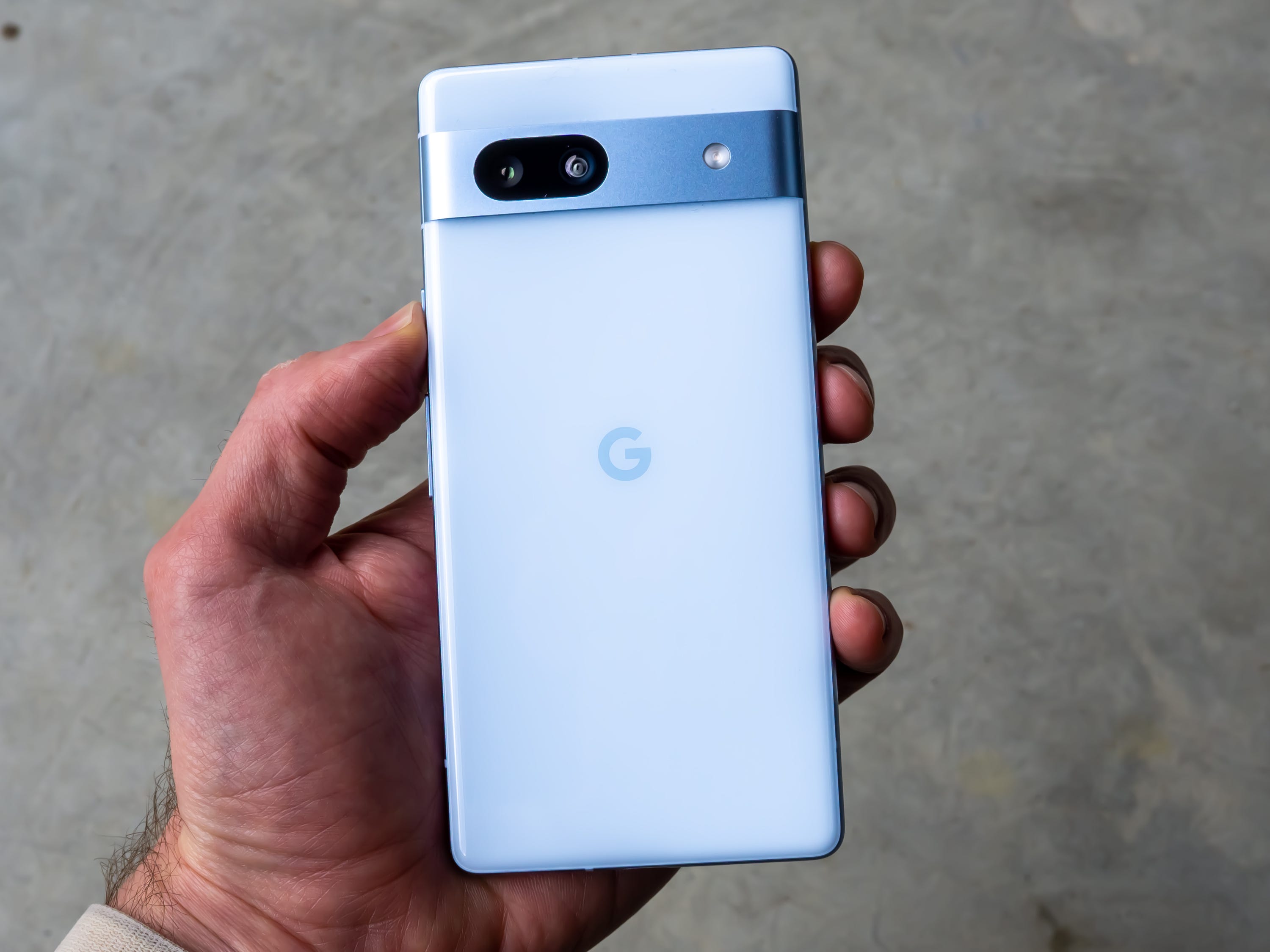 Die Rückseite des Google Pixel 7a in der Farboption Blau, in einer Hand gehalten.