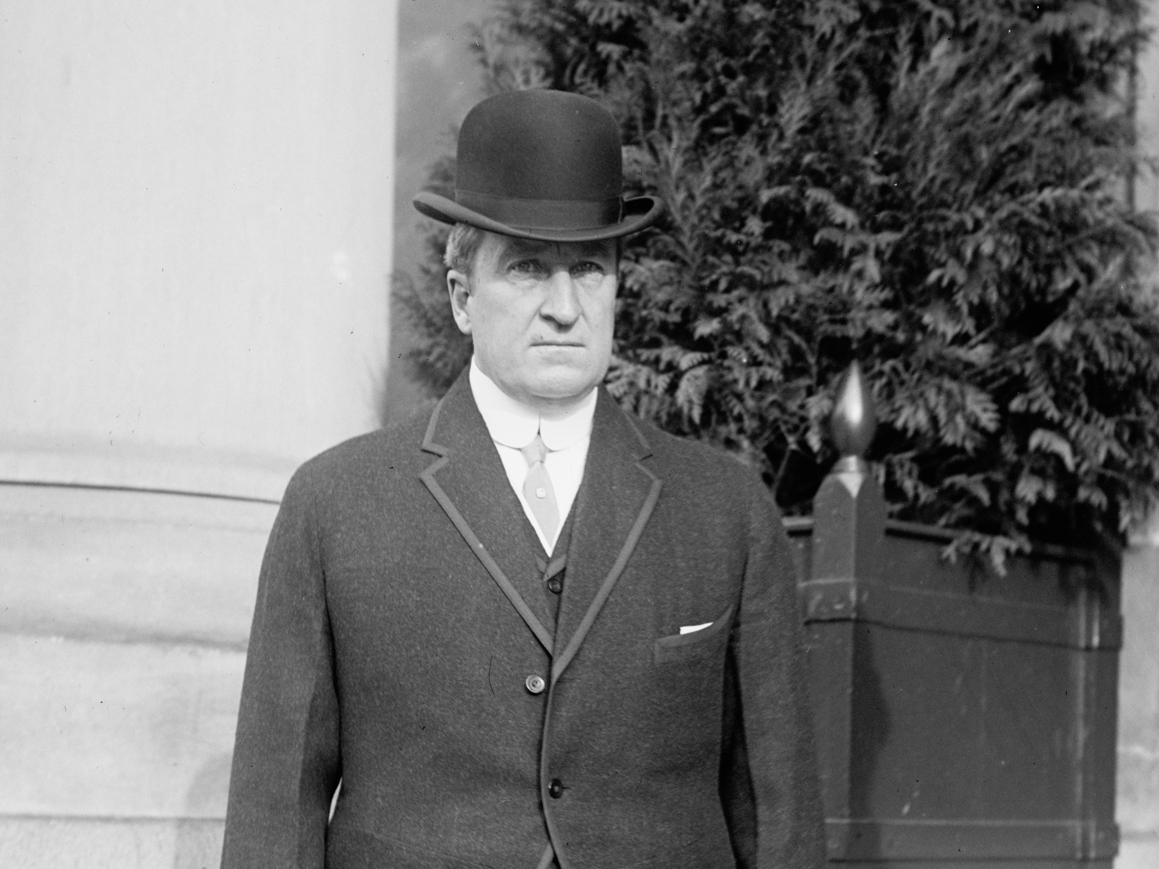 John A. Dix steht mit strengem Gesichtsausdruck vor einem Gebäude und trägt einen Hut und einen schwarzen Blazer.