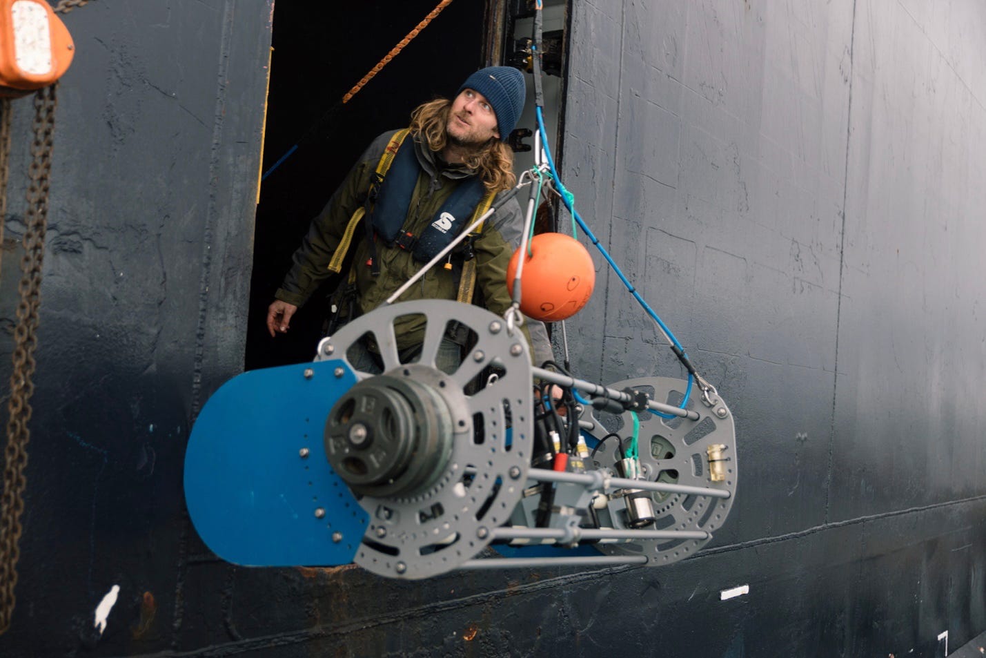 Ein Mann mit Mütze steht in einem Kreuzfahrtschiff mit einer riesigen Kameraausrüstung, die gerade ins Wasser gelassen wird