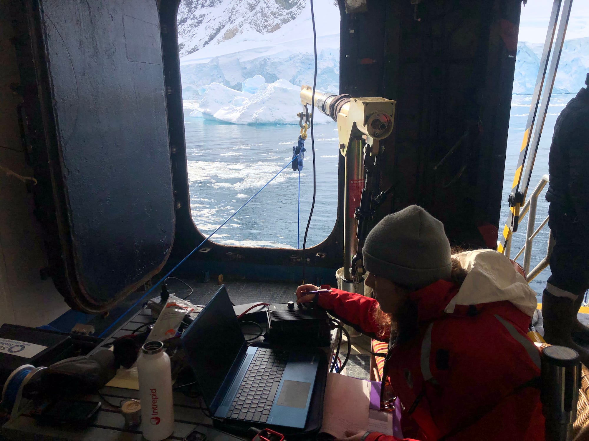 Eine Frau in einem Kreuzfahrtschiff mit einem Laptop und anderen Geräten und der antarktischen Landschaft vor dem Fenster