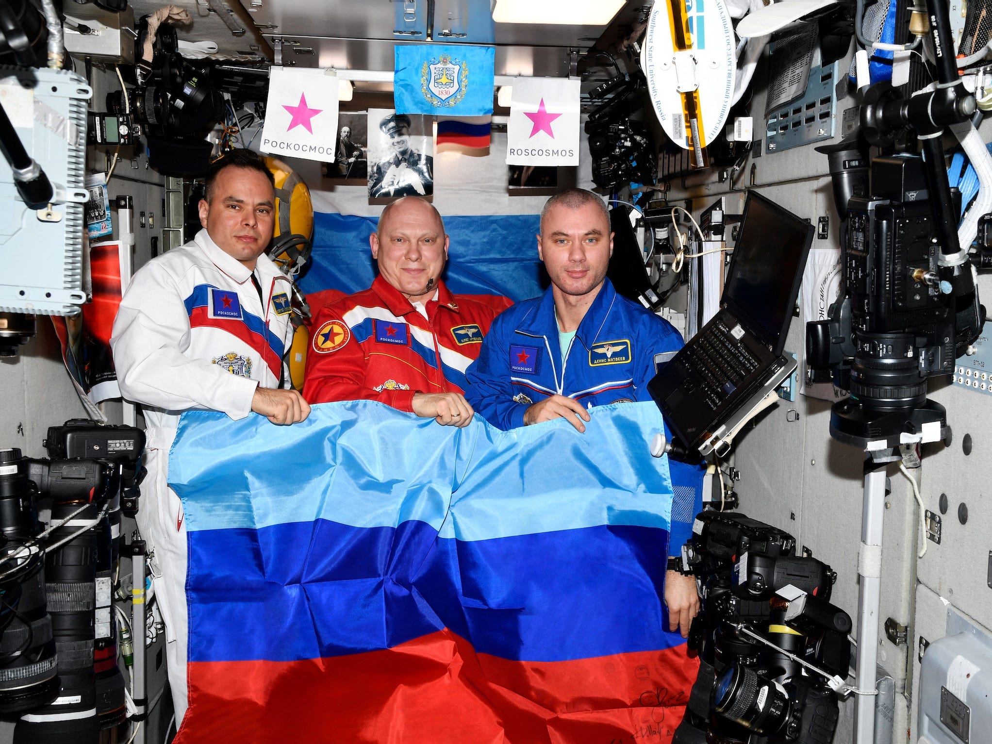 Drei Kosmonauten posieren mit blau-rot gestreifter Flagge in der Raumstation