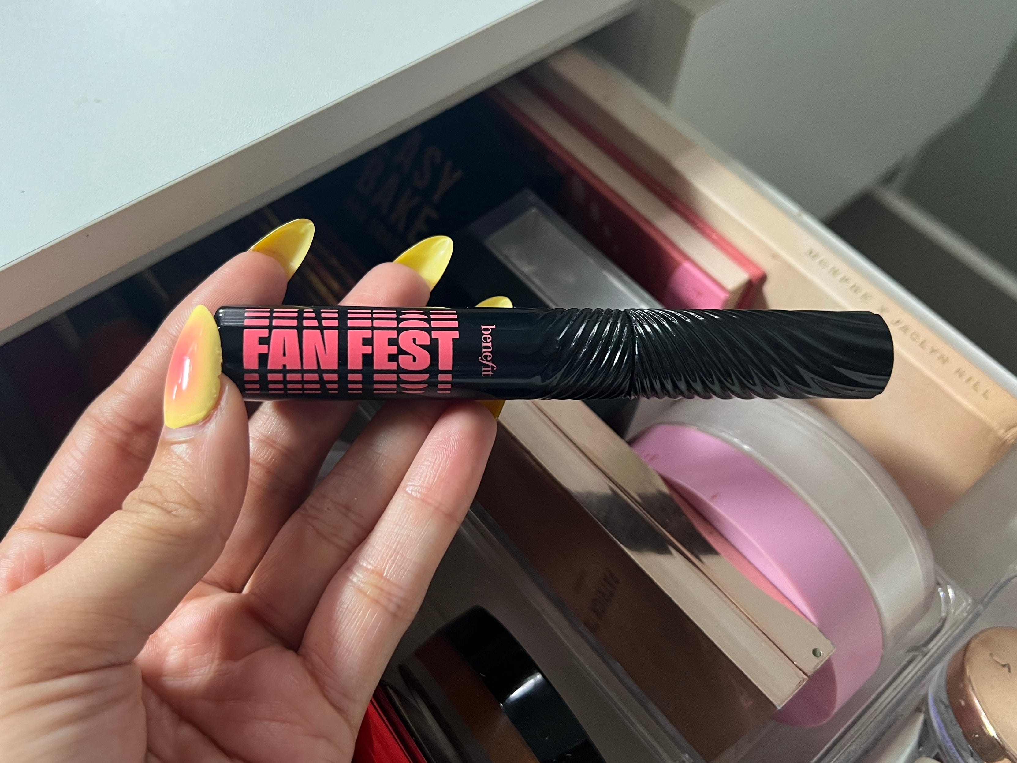 Eine Hand mit gelben Fingernägeln hält eine schwarze Tube mit der Aufschrift „Benefit Fan Fest“ in rosa Schrift vor eine offene Make-up-Schublade