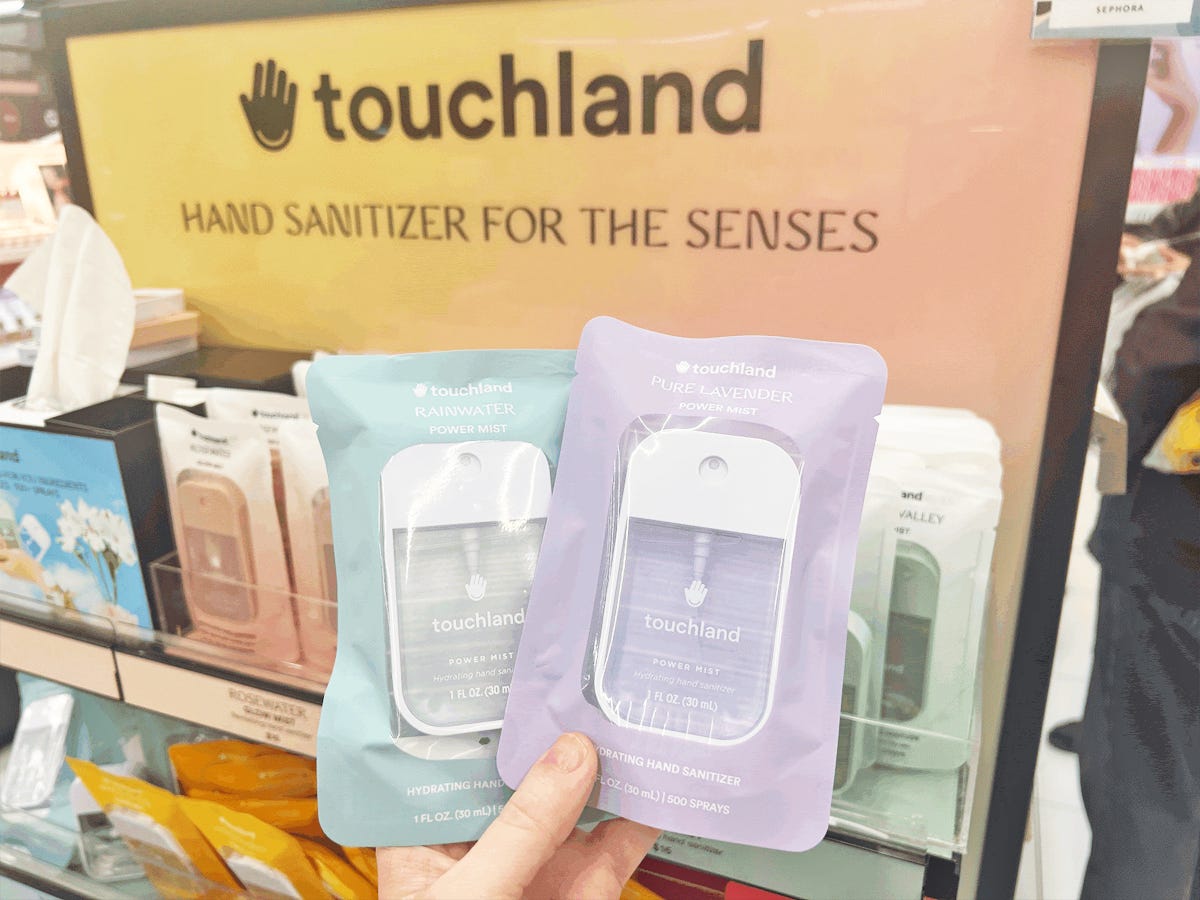 Eine Hand hält zwei Packungen Touchland-Händedesinfektionsmittel vor einem orangefarbenen Schild mit der Aufschrift „Touchland-Händedesinfektionsmittel für die Sinne“.  Eine Packung ist hellblau und die andere lila