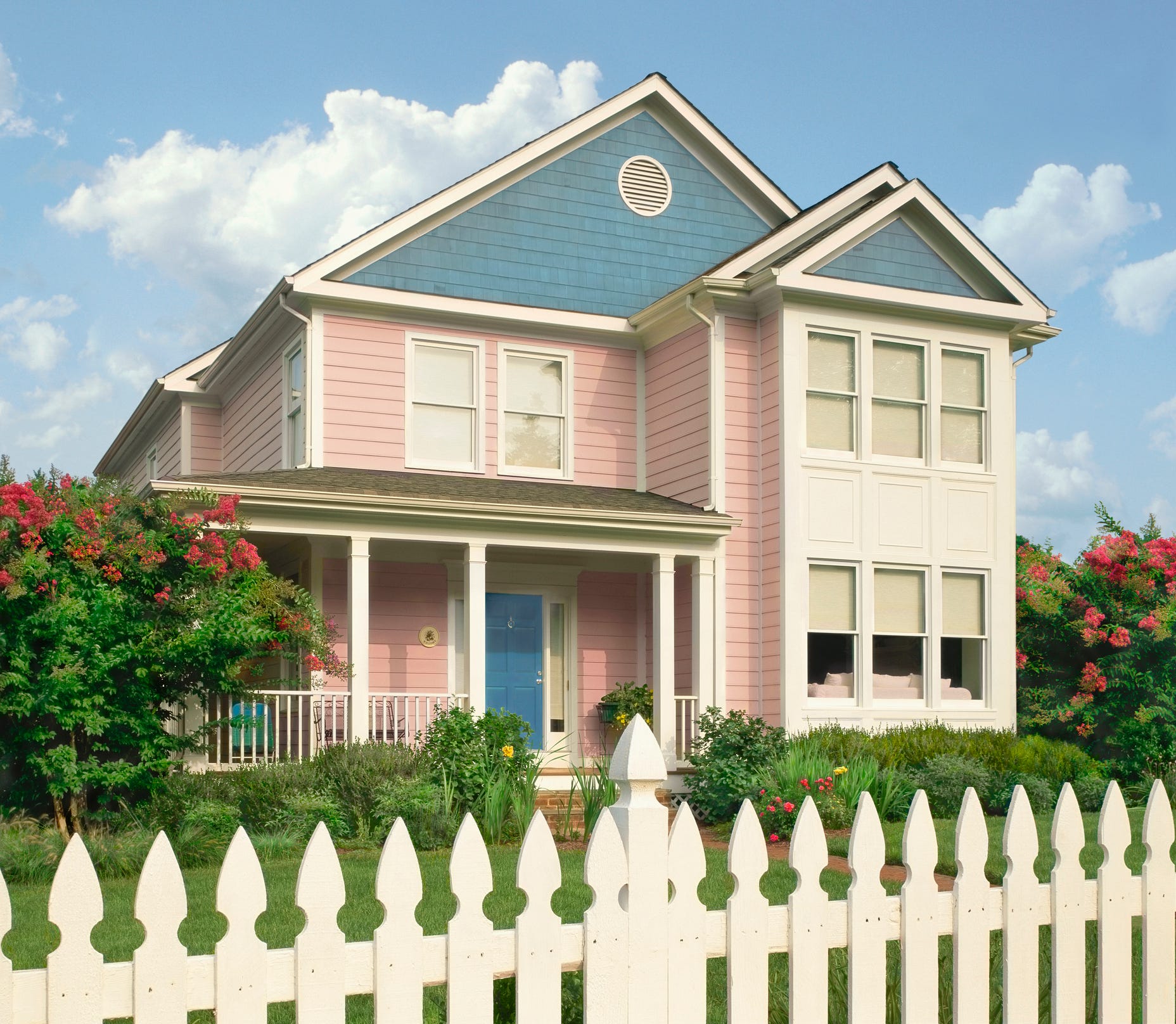 Dargestellt ist ein zweistöckiges Haus mit blassrosa und blassblauen Fassaden.  Im Vordergrund steht ein weißer Zaun.