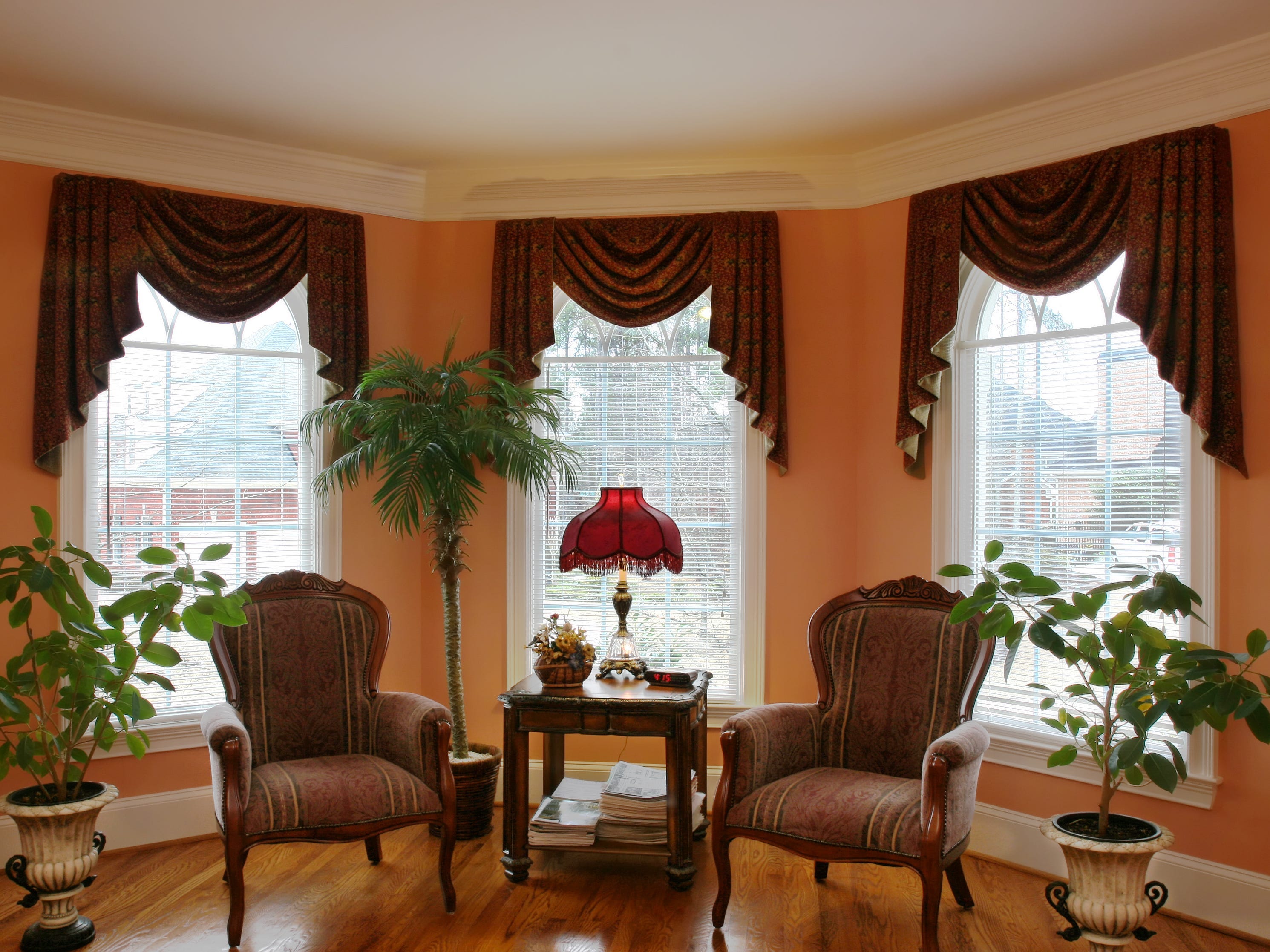 Wohnzimmer mit zwei um einen Tisch angeordneten Stühlen neben Topfpflanzen und orangefarbenen Wänden.  Dunkelrote Fenstervorhänge rahmen drei Fenster ein.