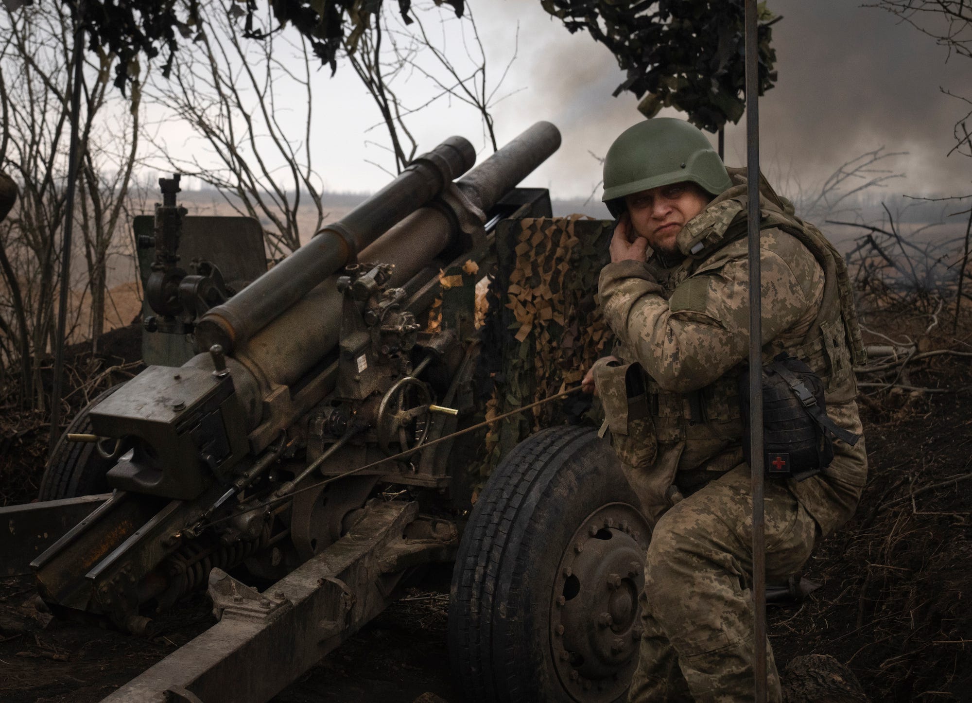 Ein ukrainischer Soldat in Kampfausrüstung und Helm hockt neben einer M101-Haubitze und hält sich das Ohr zu