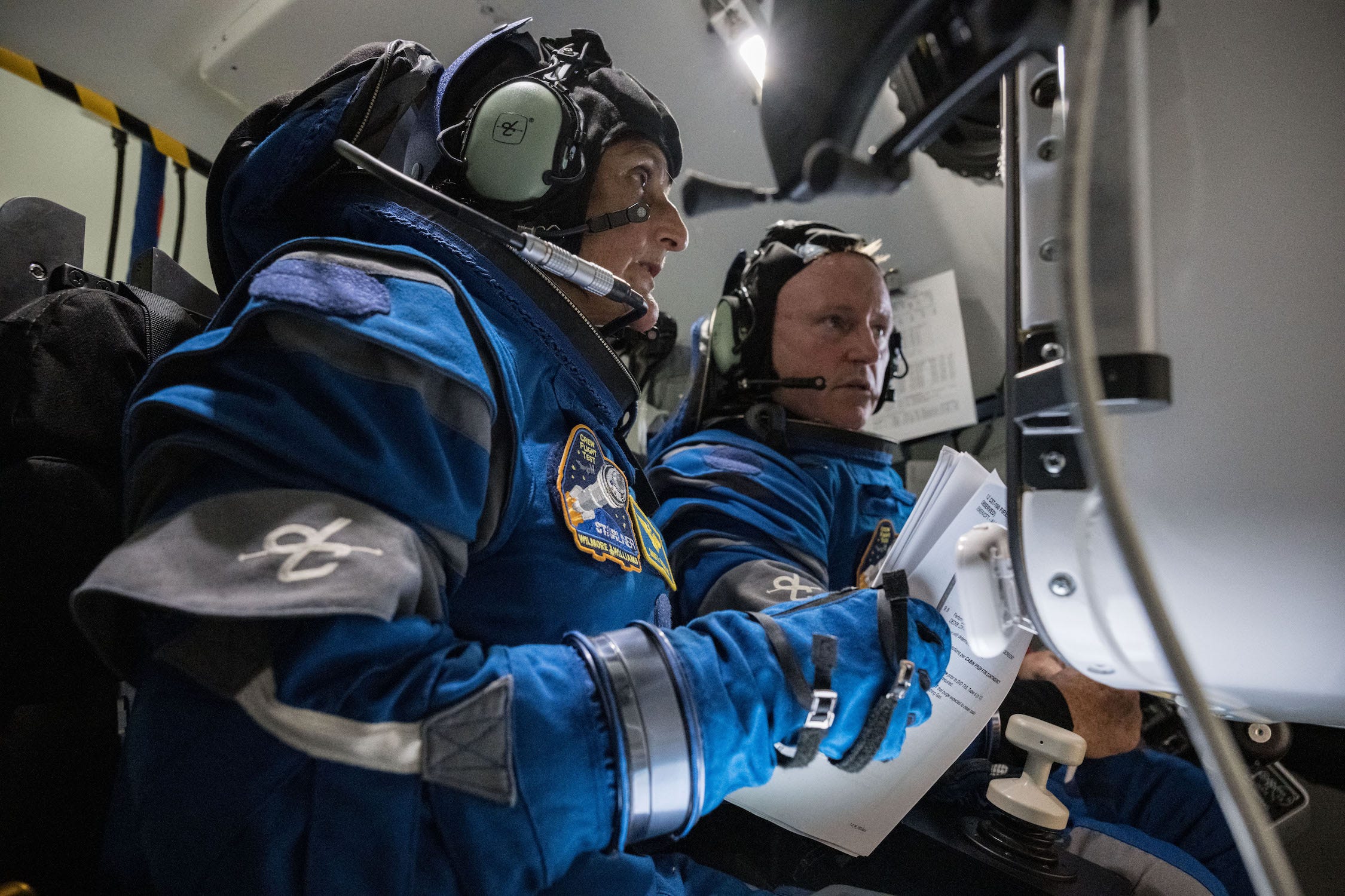 Zwei Astronauten in blauen Raumanzügen in einem Raumschiff halten Papiere in der Hand und schauen auf ein Armaturenbrett