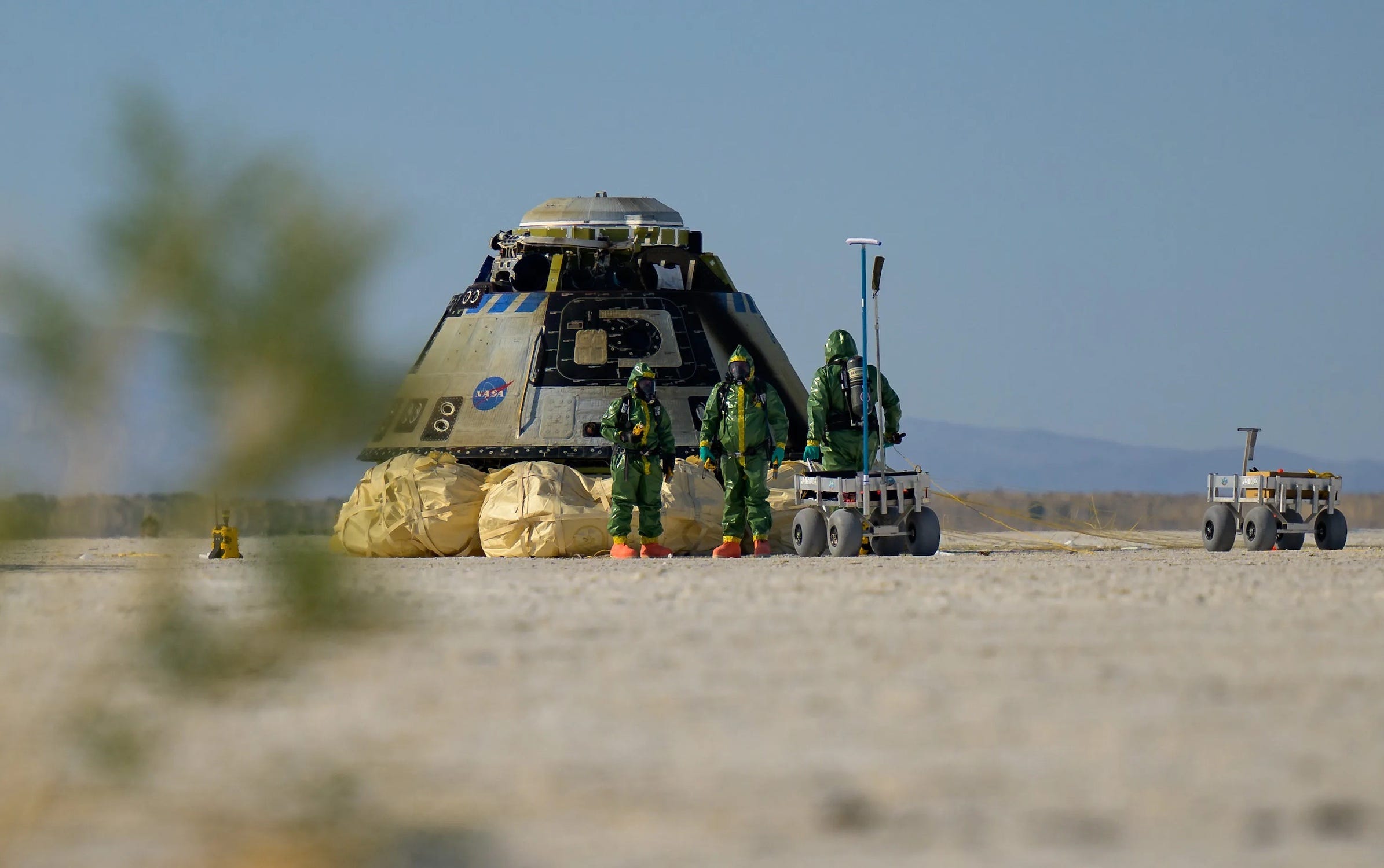 Drei Menschen in grünen Schutzanzügen stehen neben einer Raumkapsel, die auf großen Airbags auf zwei metallenen Rover-ähnlichen Karren in der Wüste sitzt