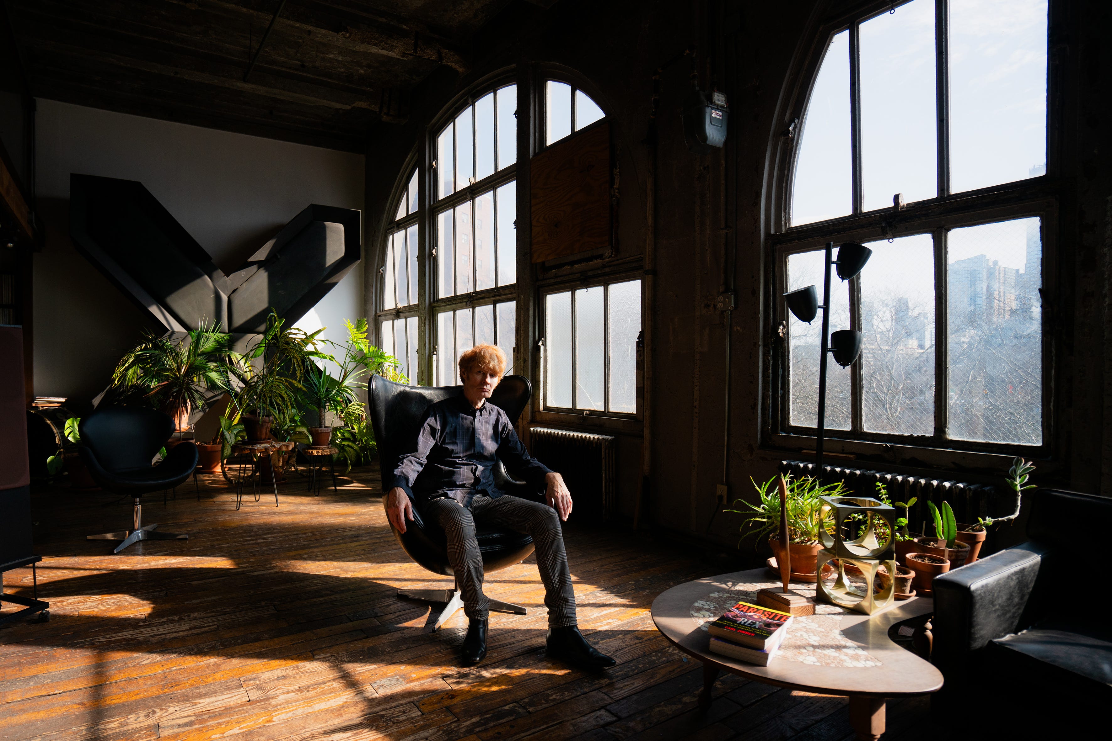 JG Thirwell sitzt in einem modernen Stuhl, umgeben von Pflanzen und riesigen Fenstern in seinem Loft.