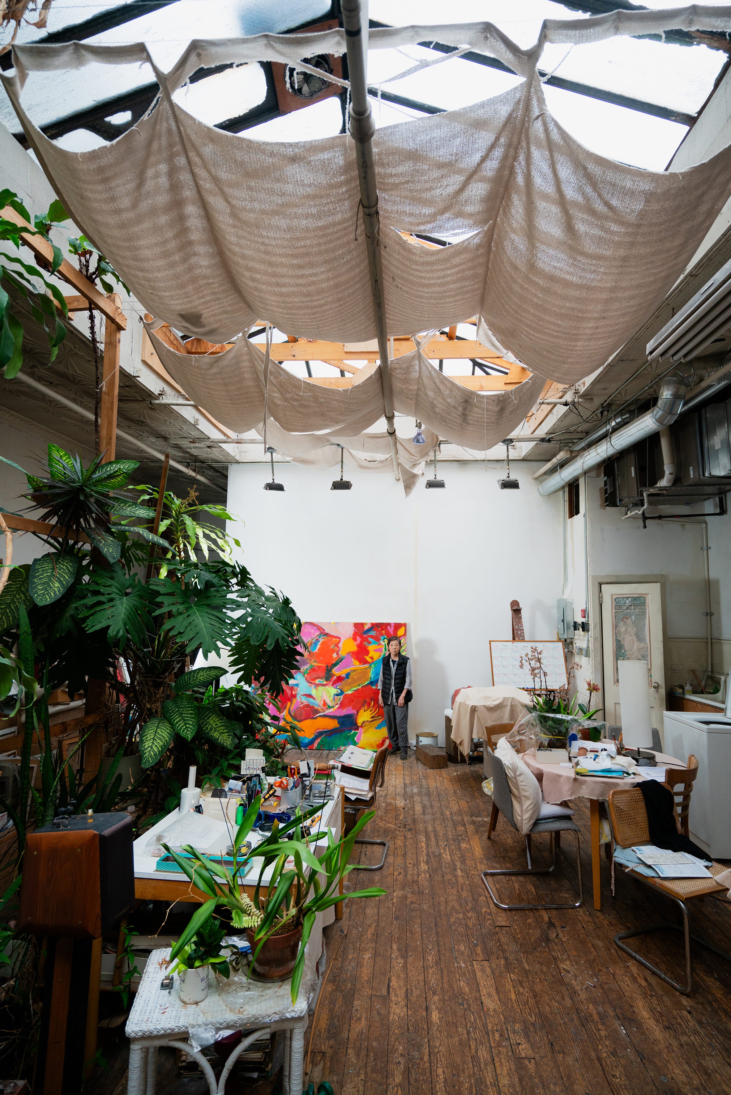 Kimiko Fujimura steht in einiger Entfernung in ihrem Chinatown-Studio mit großen grünen Pflanzen und Technicolor-Gemälden.