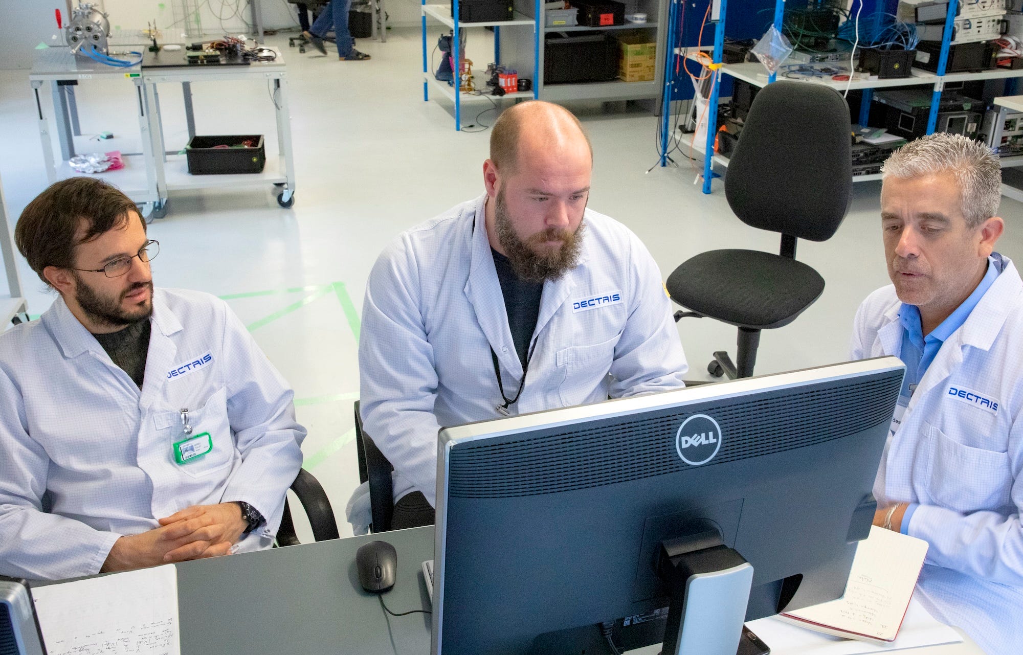 Drei Männer in weißen Laborkitteln sitzen an einem Schreibtisch und schauen auf einen Computer
