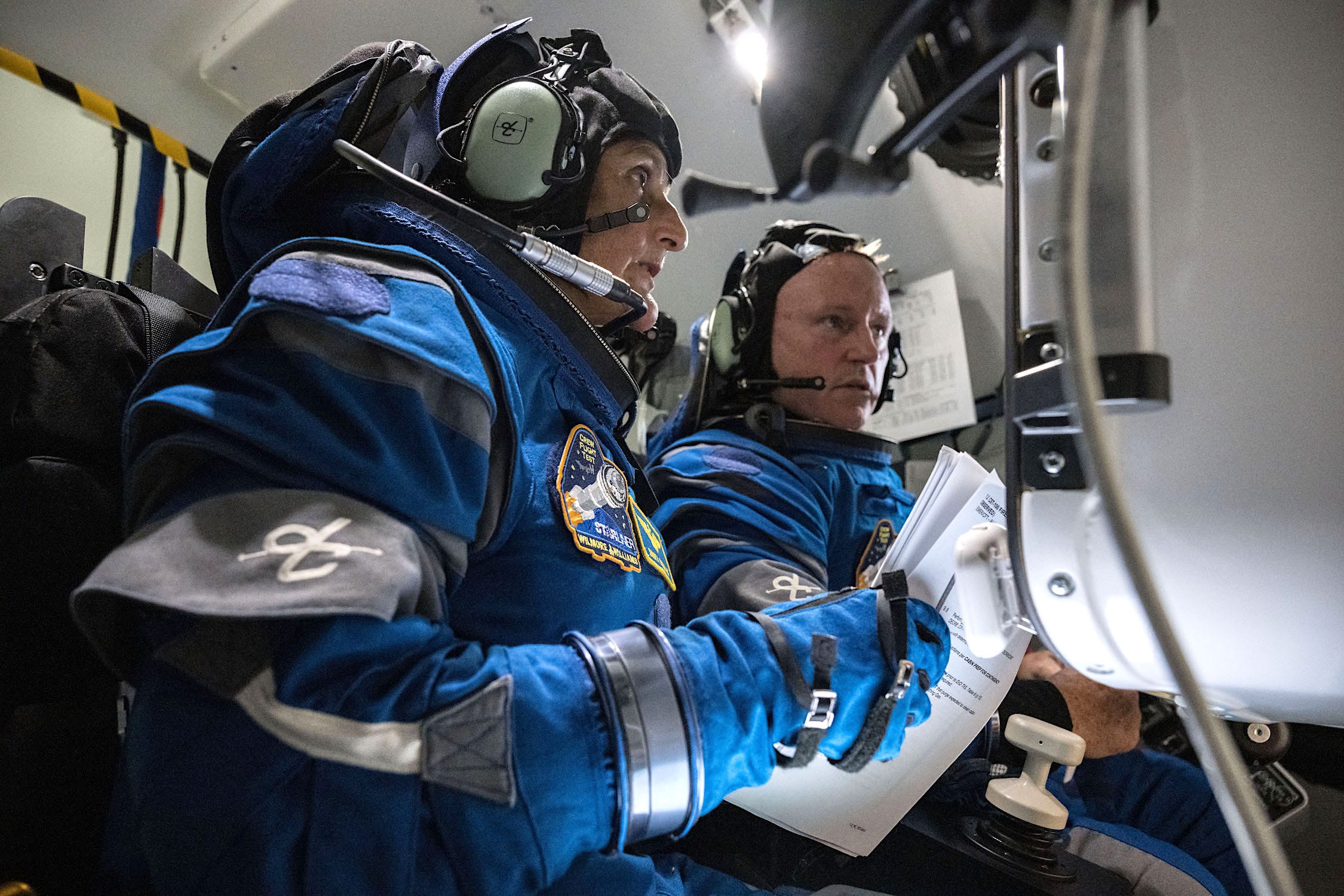 Zwei Astronauten in blauen Raumanzügen in einem Raumschiff halten Papiere in der Hand und schauen auf ein Armaturenbrett
