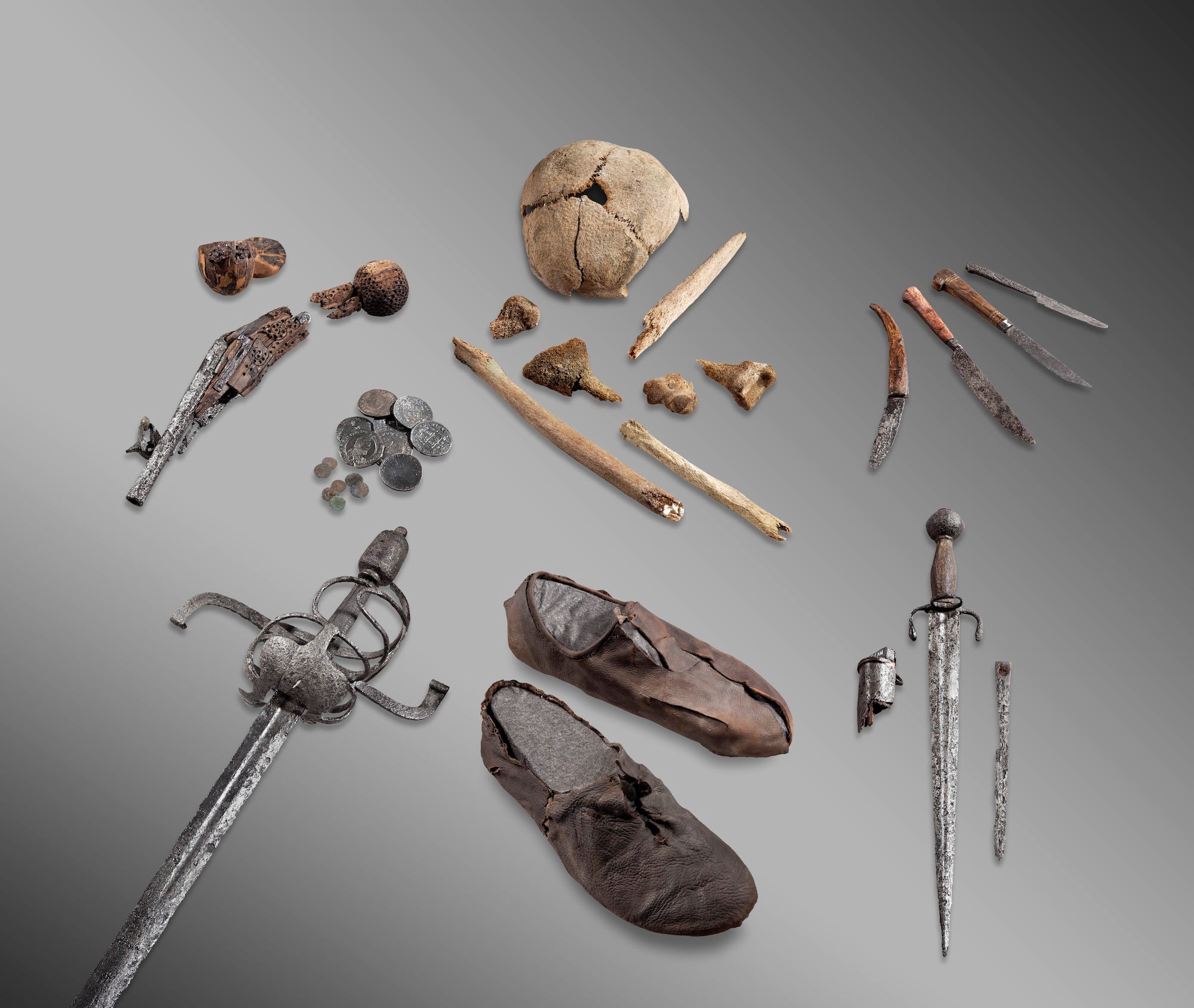 Schädelkappe, kleine Knochen, rostige Messer, Dolch, Schwert, Münzen, kaputte Pistole und abgenutzte Lederschuhe, ausgebreitet auf grauem Hintergrund