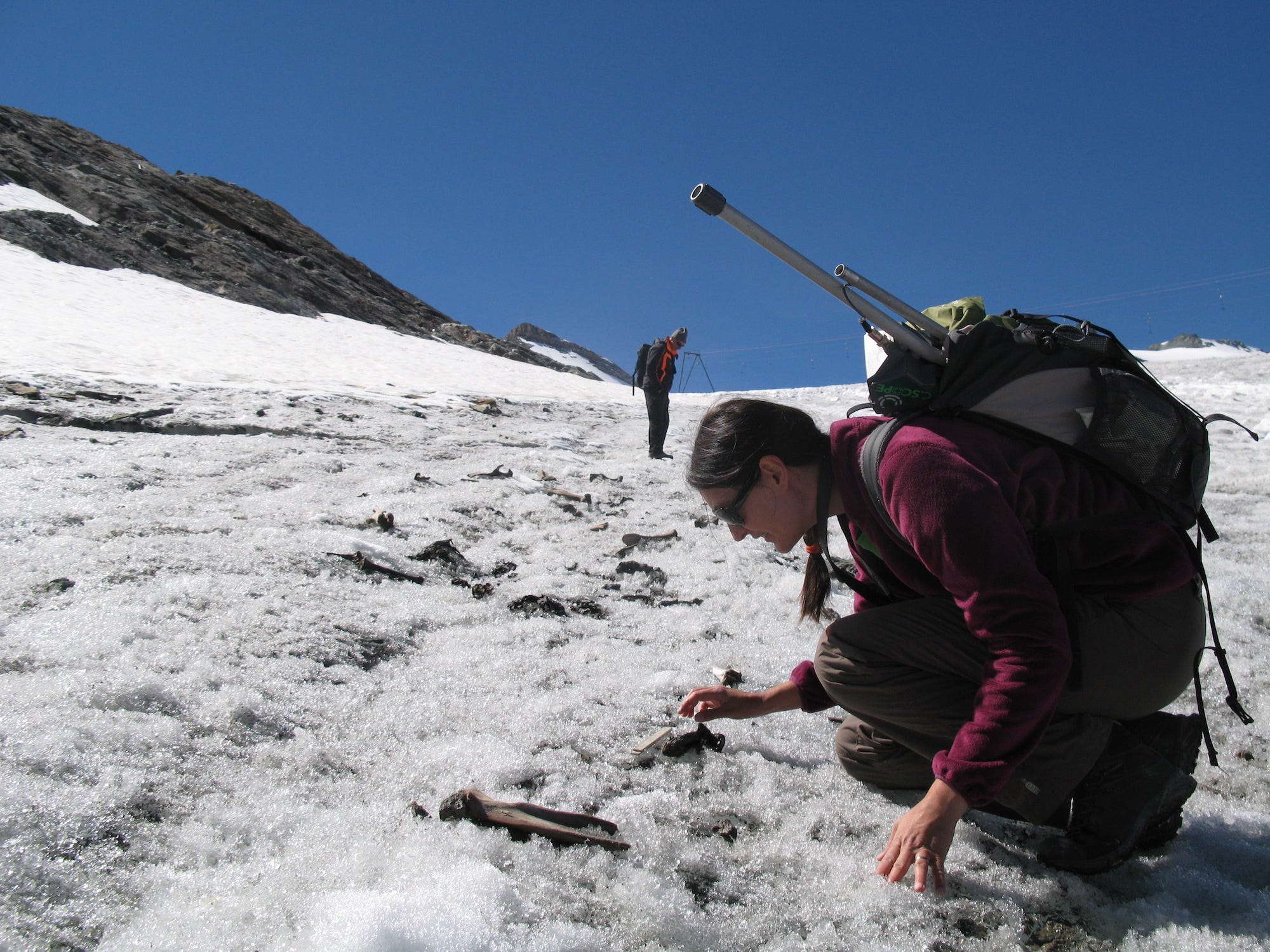 Eine Frau mit einem großen Rucksack und Stangen, die oben herausragen, hockt auf knusprigem, strukturiertem Eis und blickt auf einen Knochen, der auf dem Boden liegt, mit Berggipfeln im Hintergrund