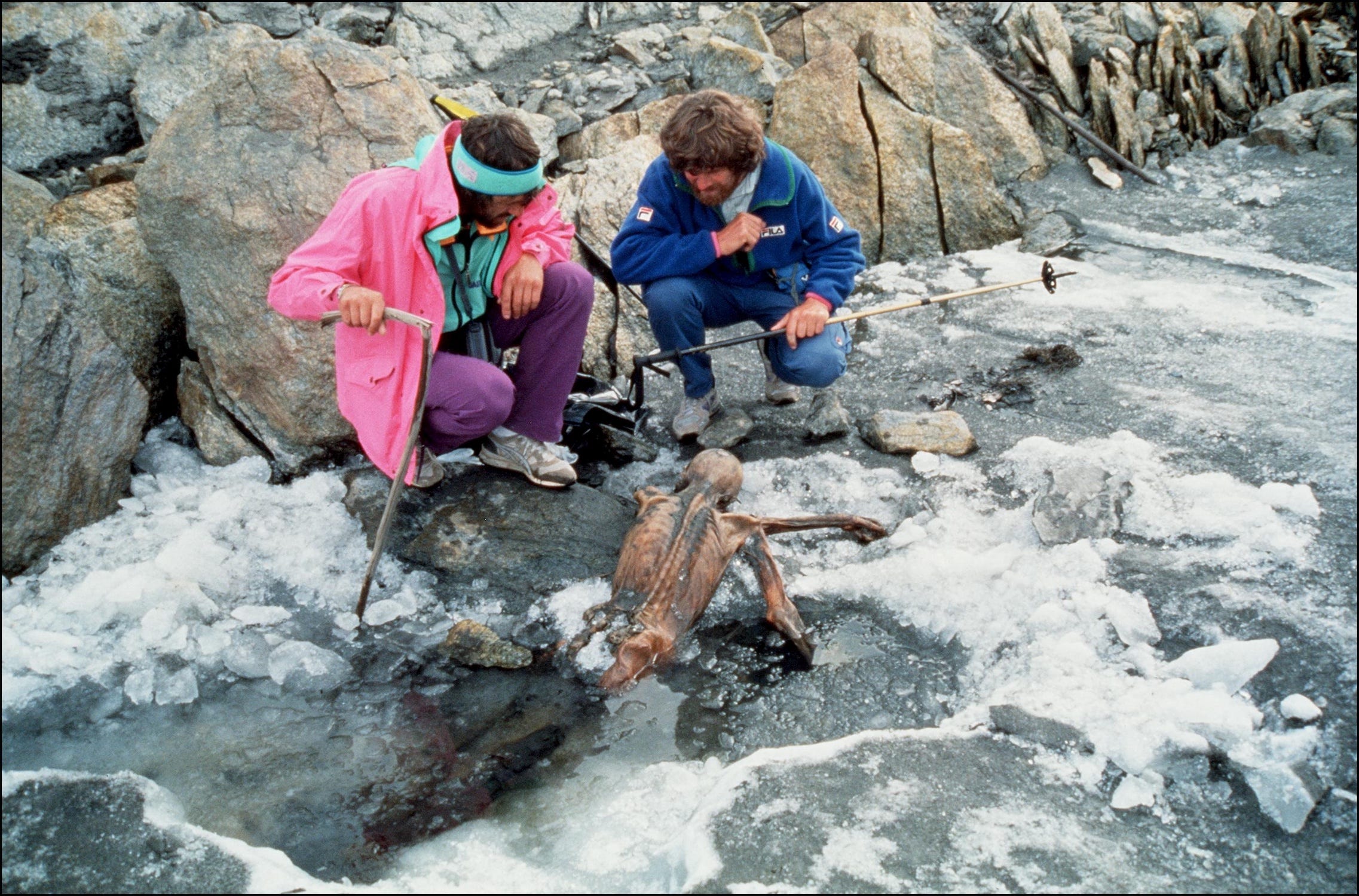 Zwei Männer mit struppigen Haaren in Wanderkleidung aus den 90er-Jahren krabbeln auf schmelzendem Eis neben einer verdeckten Mumie, die so positioniert ist, als würde sie aus einer Pfütze im Eis kriechen