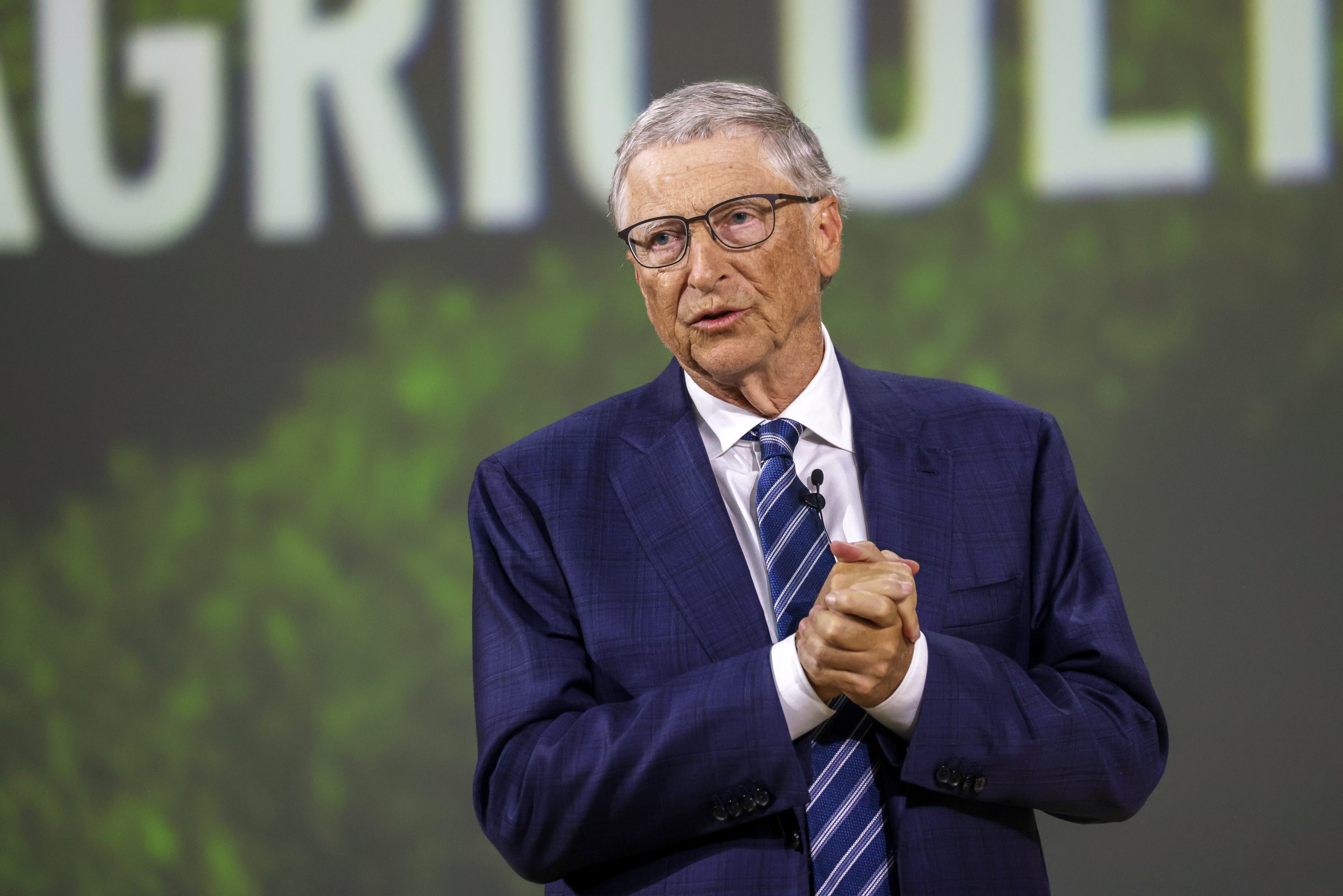 Bill Gates trägt einen dunkelblauen Anzug mit gefalteten Händen