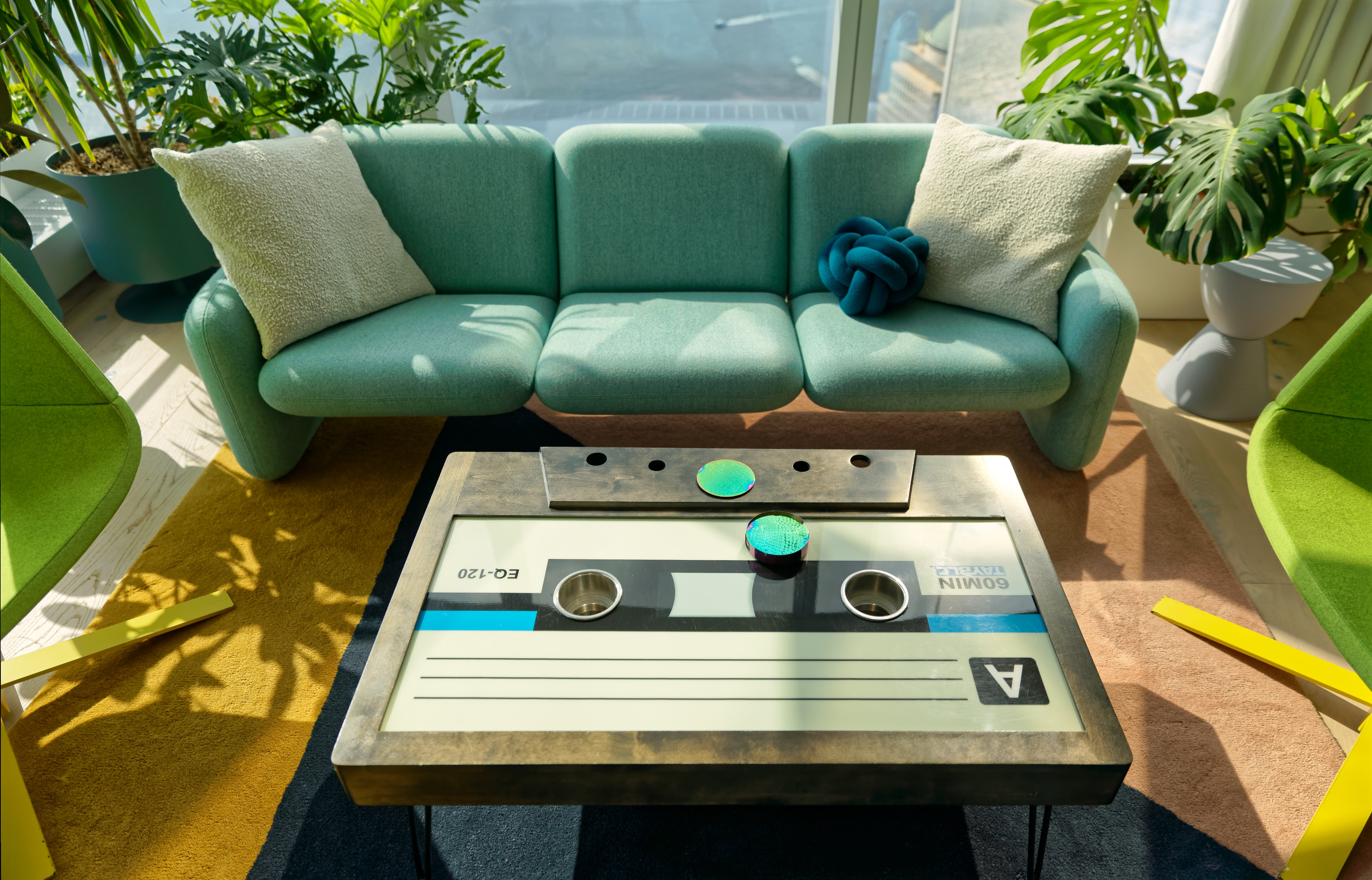Eine grüne Couch mit einem Couchtisch, der so geformt und gestaltet ist, dass er wie eine Kassette aussieht.  Der Raum hat viel natürliches Licht und ein paar Pflanzen darin.