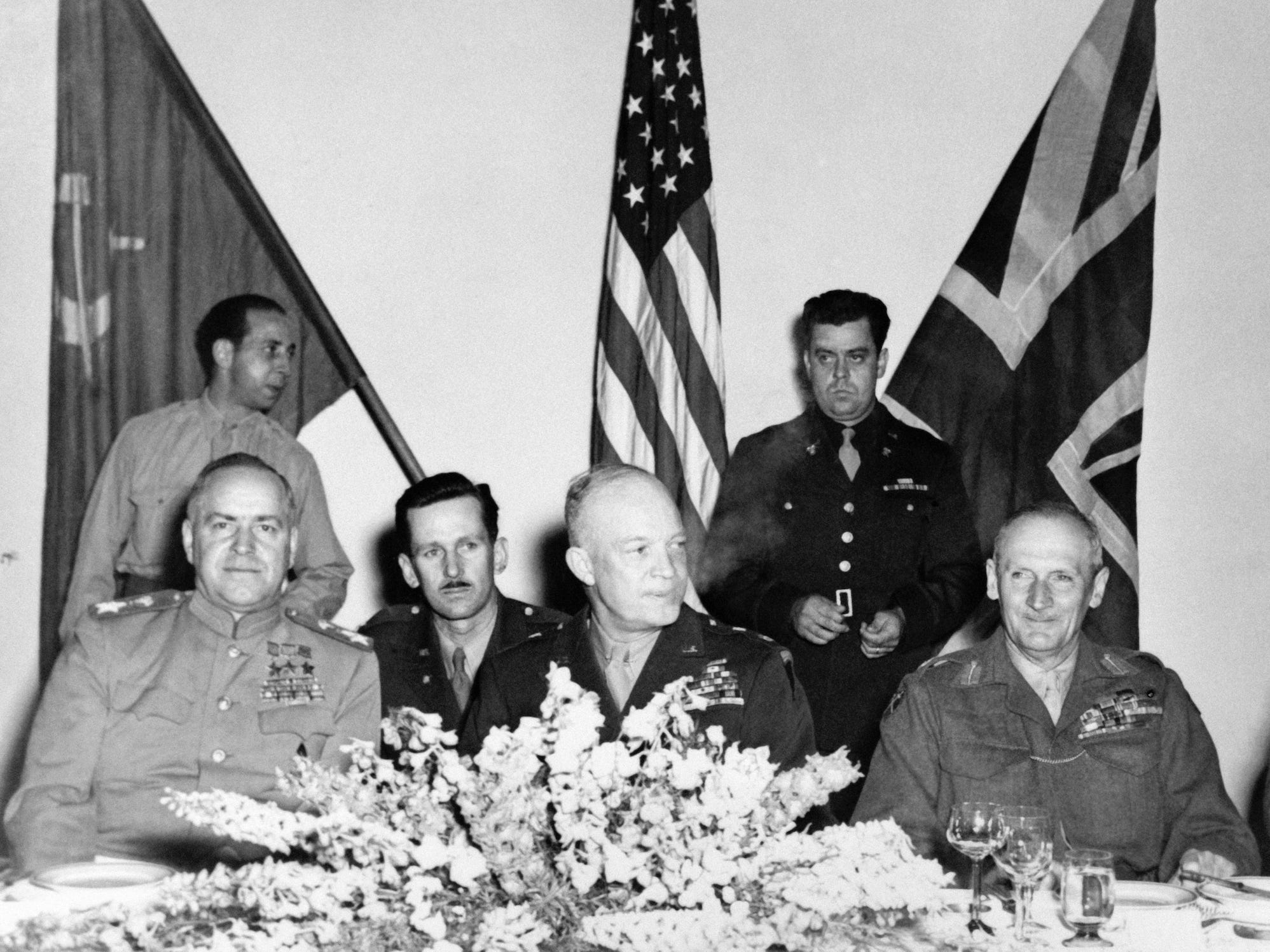 Die Generäle Bernard Montgomery, Dwight Eisenhower und Georgi Schukow sitzen an einem Tisch mit Fahnen im Hintergrund und einem Blumenarrangement davor