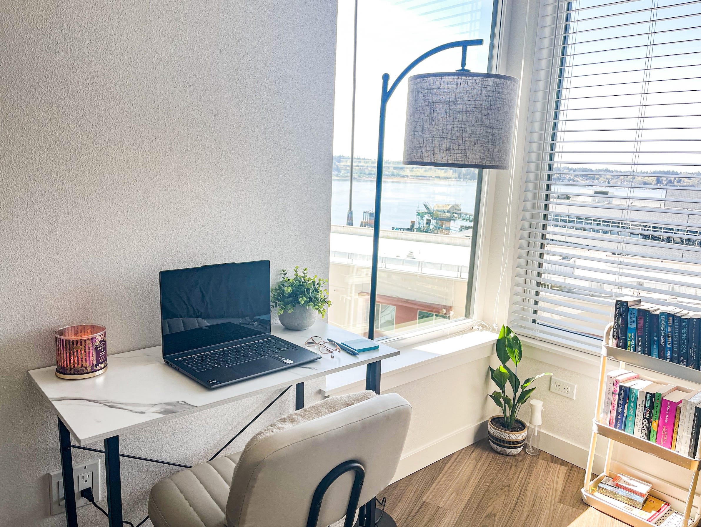 Ein Arbeitsbereich mit einem weißen Schreibtisch, einem Stuhl und einer Lampe mit Fenstern und einem Bücherregal im Hintergrund.  Auf dem Schreibtisch stehen eine Kerze und ein Laptop