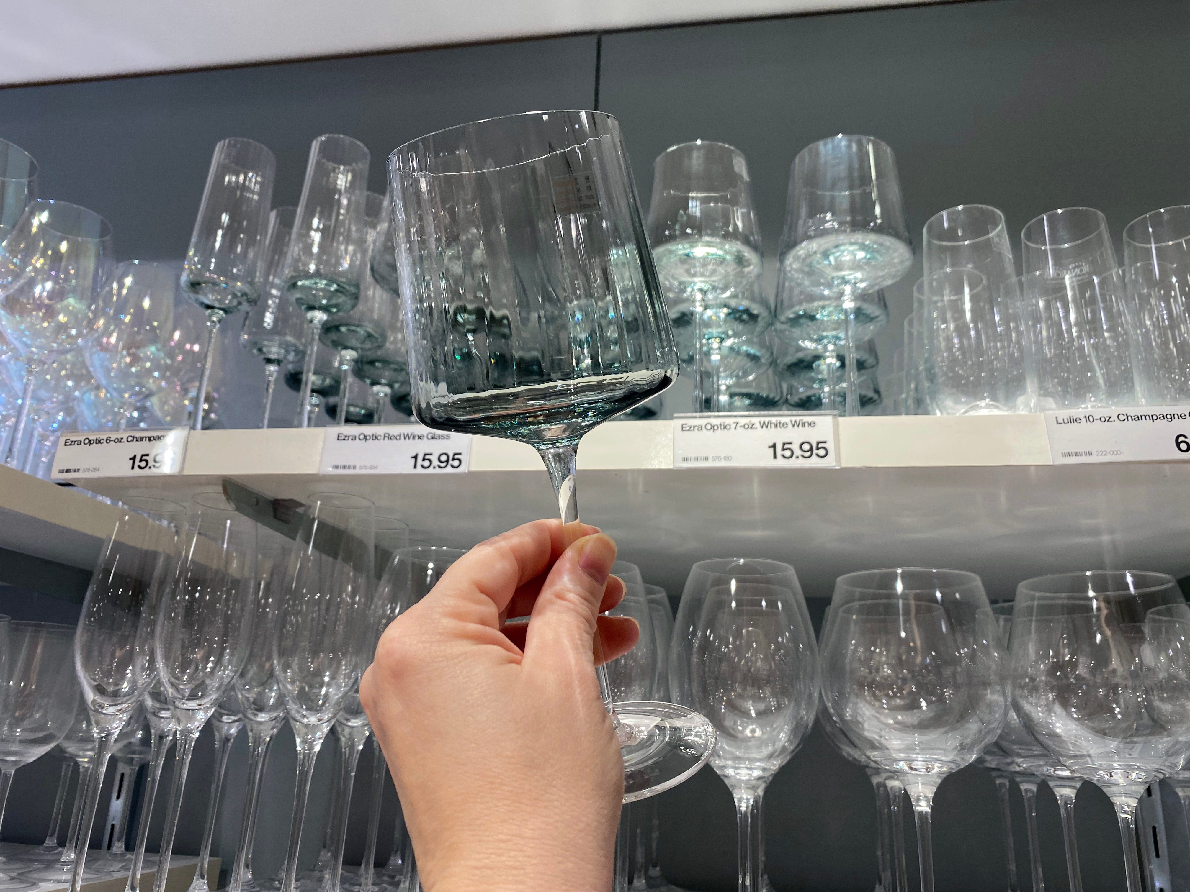 Eine Hand hält ein Trinkglas aus klarem Glas vor einem Regal mit mehreren Weingläsern. Das Glas im Vordergrund hat einen langen Stiel und eine rechteckige Form
