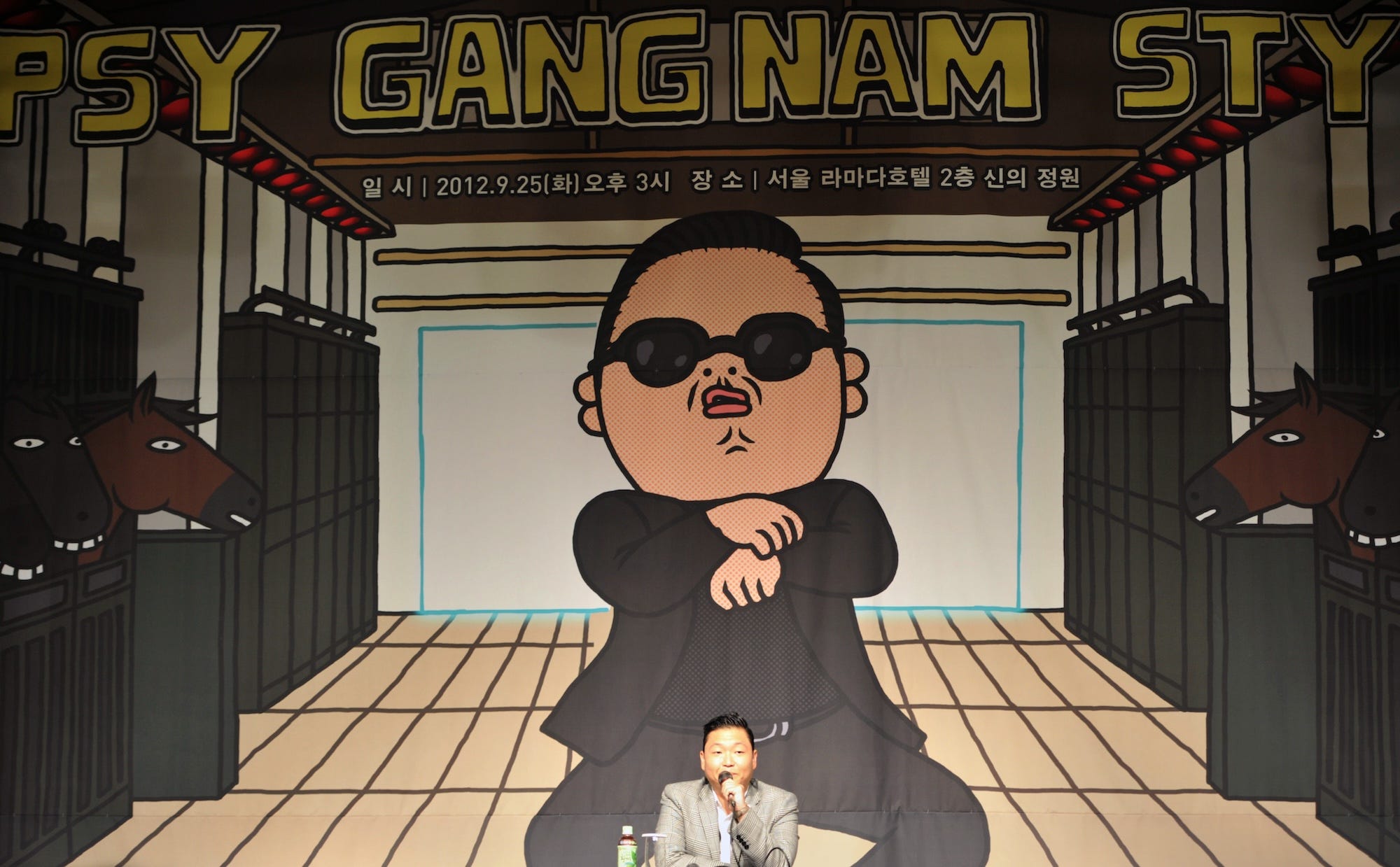 Der südkoreanische Popstar Psy spricht in ein Mikrofon vor einer riesigen Kulisse, auf der eine Cartoon-Version von ihm selbst zu sehen ist, wie er den Gangnam-Style-Tanz aufführt.