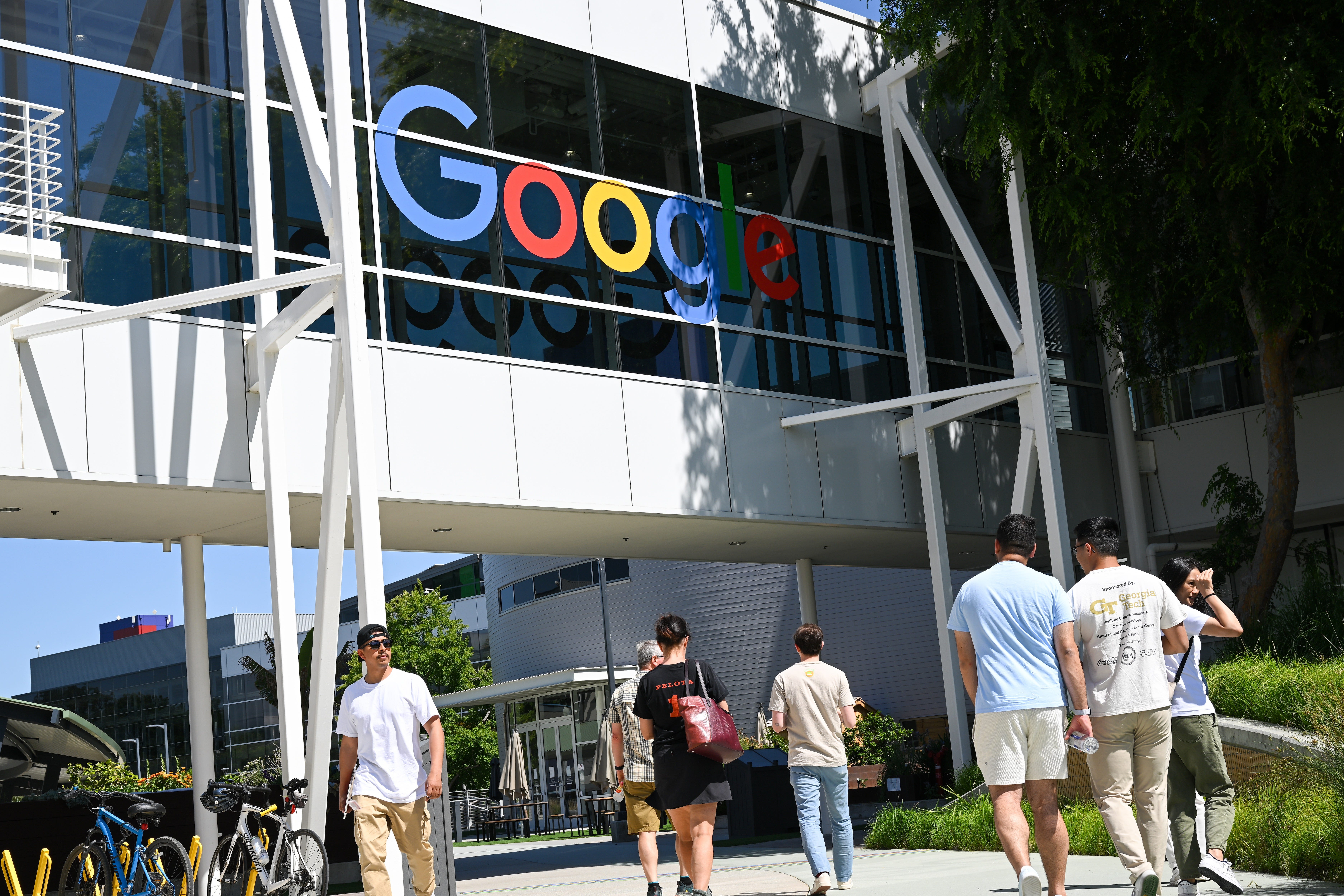 Google-Mitarbeiter gehen auf dem Googleplex-Campus unter einer Brücke mit dem Google-Logo hindurch.
