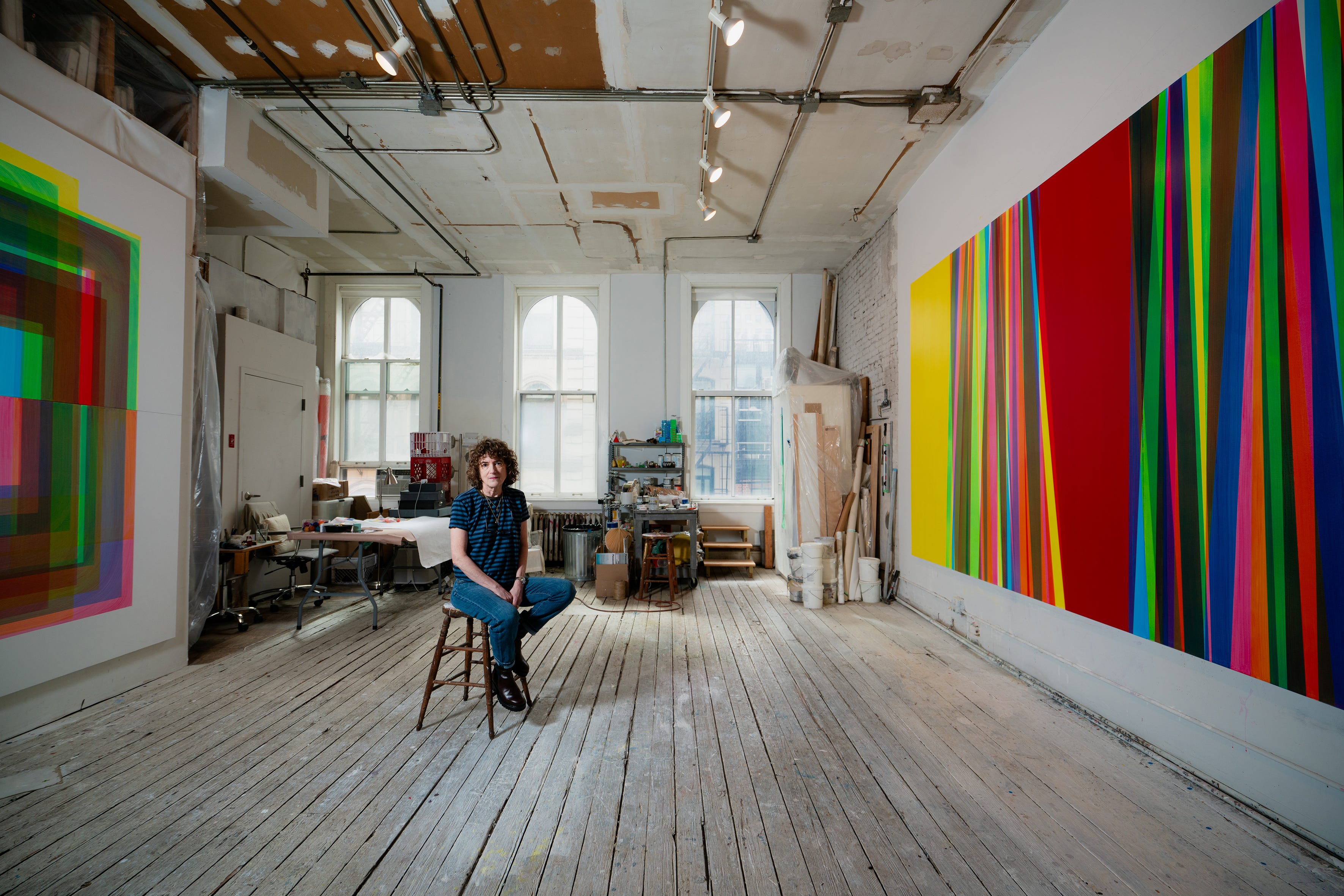 Betsy Kaufman sitzt in ihrem industriellen Atelier mit regenbogenfarbenen Gemälden an der Wand.