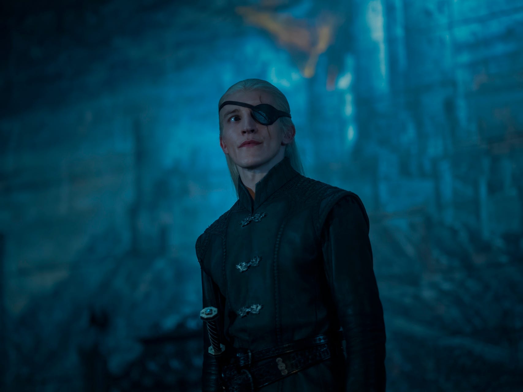 Ewan Mitchell als Aemond Targaryen, trägt schwarze Kleidung, ein Schwert an der Hüfte und eine Augenklappe über dem linken Auge. Er hat langes blondes Haar, das halb nach hinten gebunden ist.