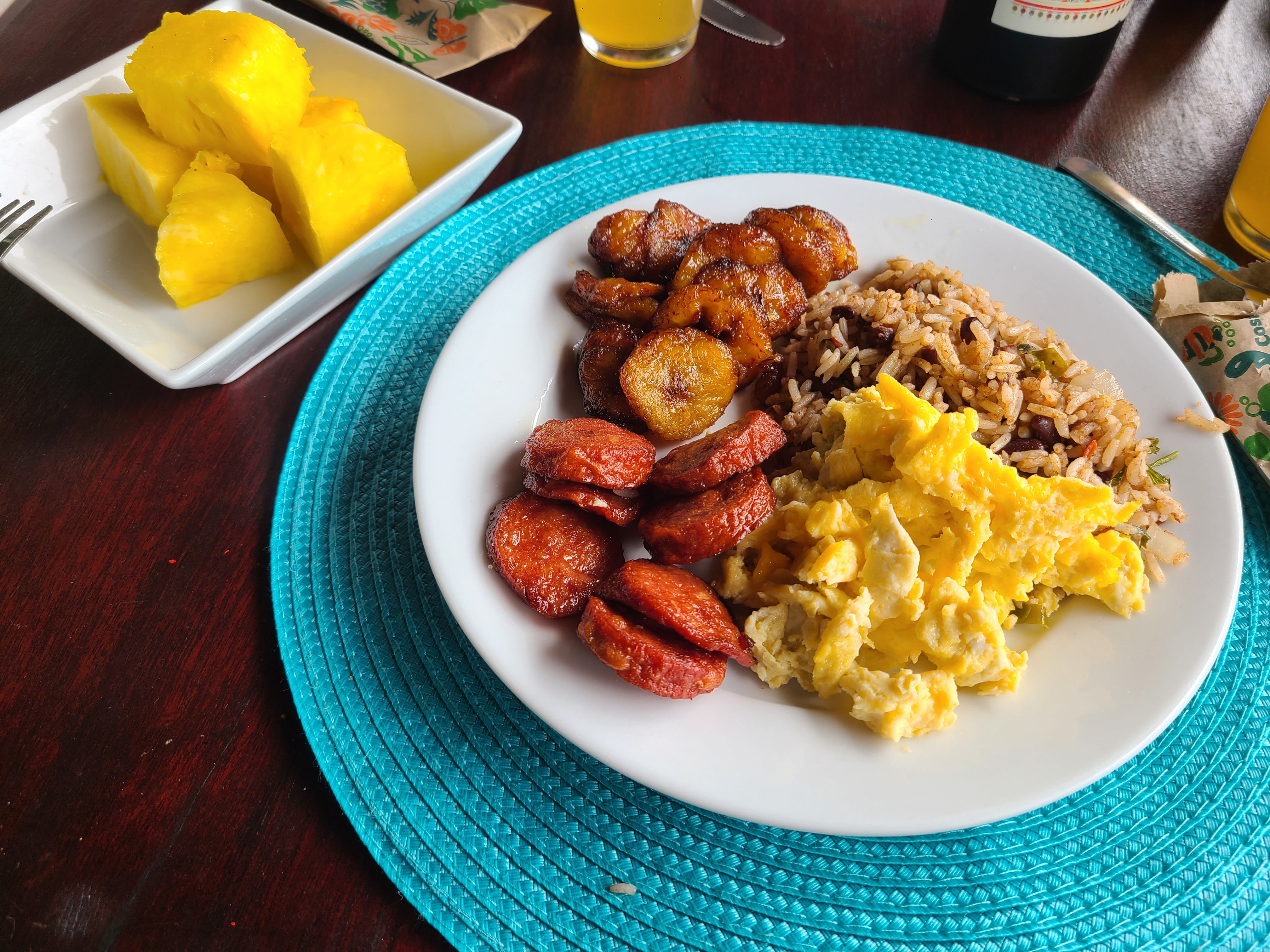 Ein weißer Teller mit einem typischen costa-ricanischen Frühstück: Eier, Gallo Pinto, Wurst, Kochbananen und Ananas.
