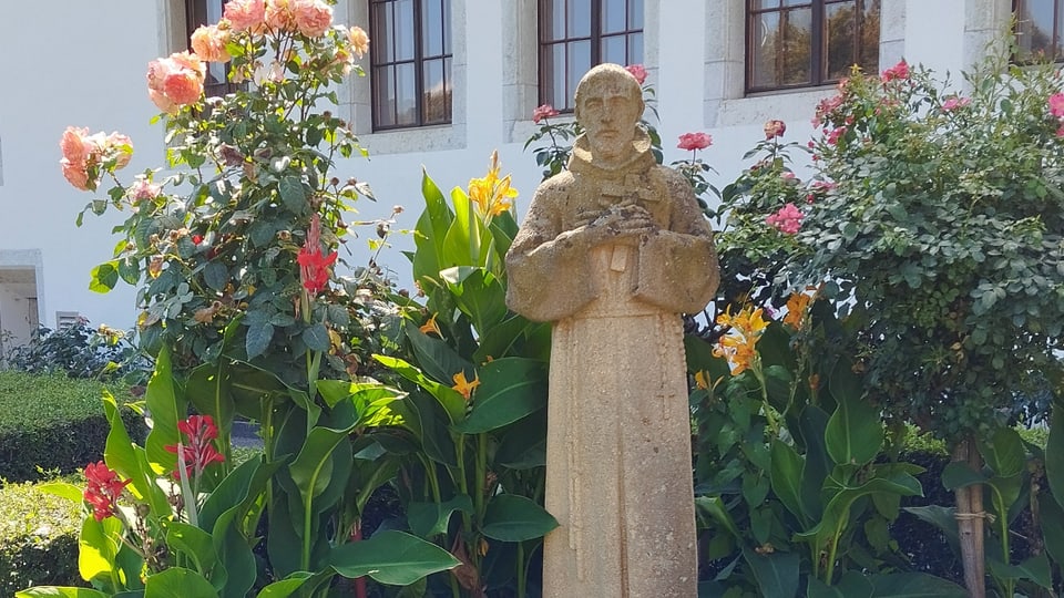 Statue of a monk surrounded by colorful flowers in front of a building.