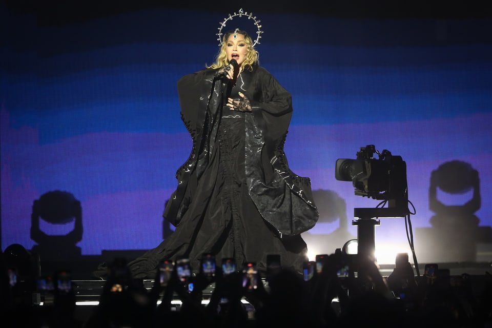 Singer in black outfit and headdress on stage in front of audience with raised cell phones.