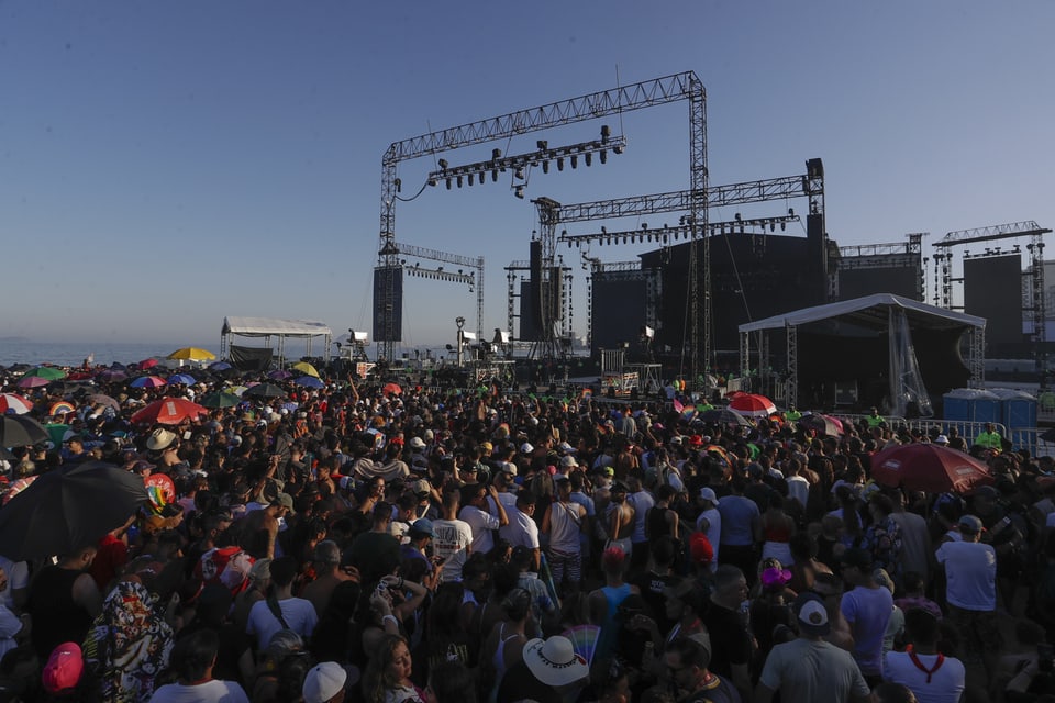 Crowd at an open-air music festival with a large stage and parasols.