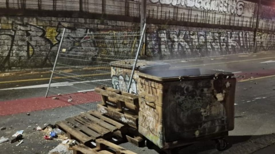 Garbage containers and pallets on a street with graffiti wall in the background.