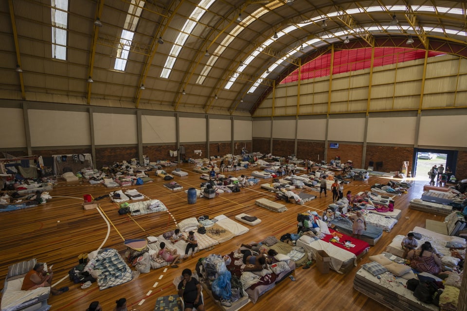 An emergency shelter in a sports hall with a curved roof.  People lie on flat mattresses on the floor.