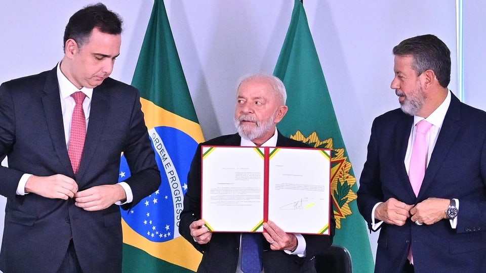Brazilian President Luiz Inácio Lula holds a document in the air, two politicians stand next to him.