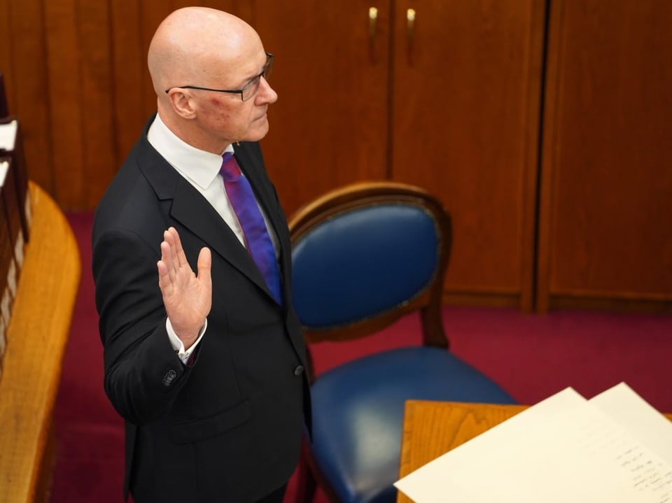 Bald man in suit taking oath near document on desk in courtroom.