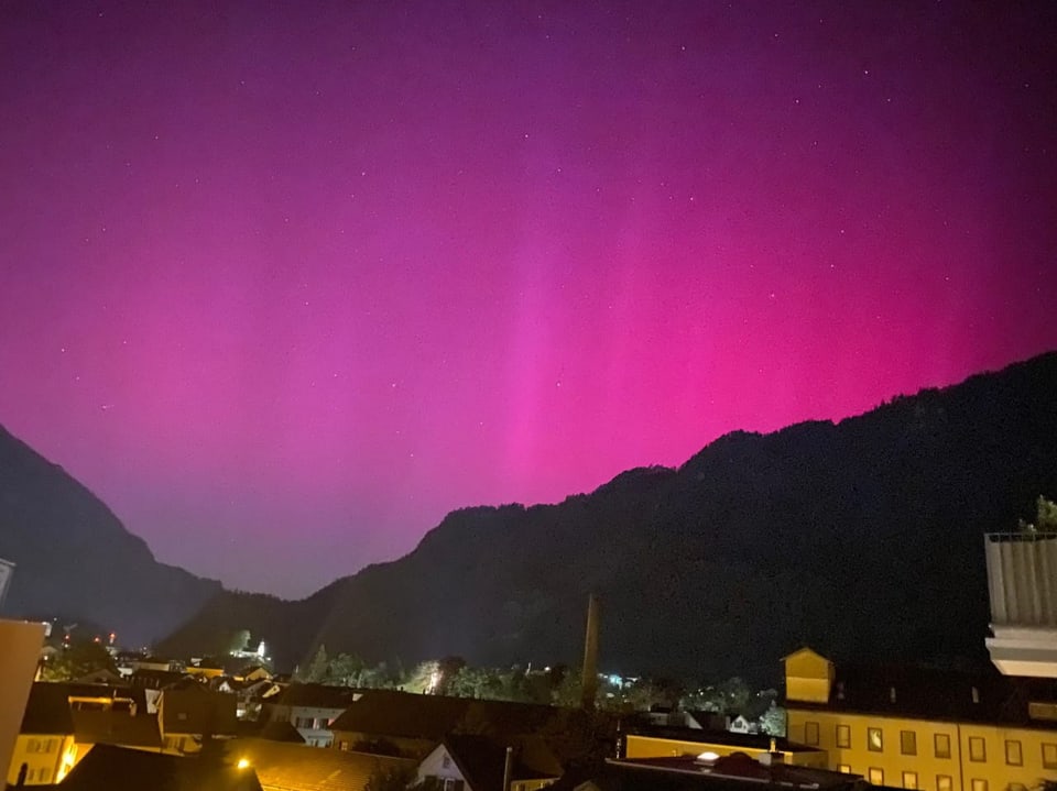 Night view of a city with pink northern lights over mountains.