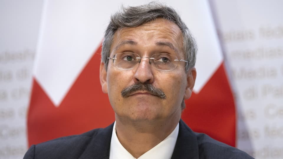 Middle-aged man with mustache in suit in front of two flags