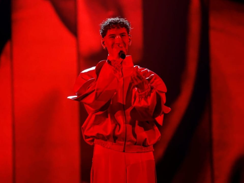 Sänger in rotem Outfit performt auf einer Bühne vor einem mikrofonierten Hintergrund.