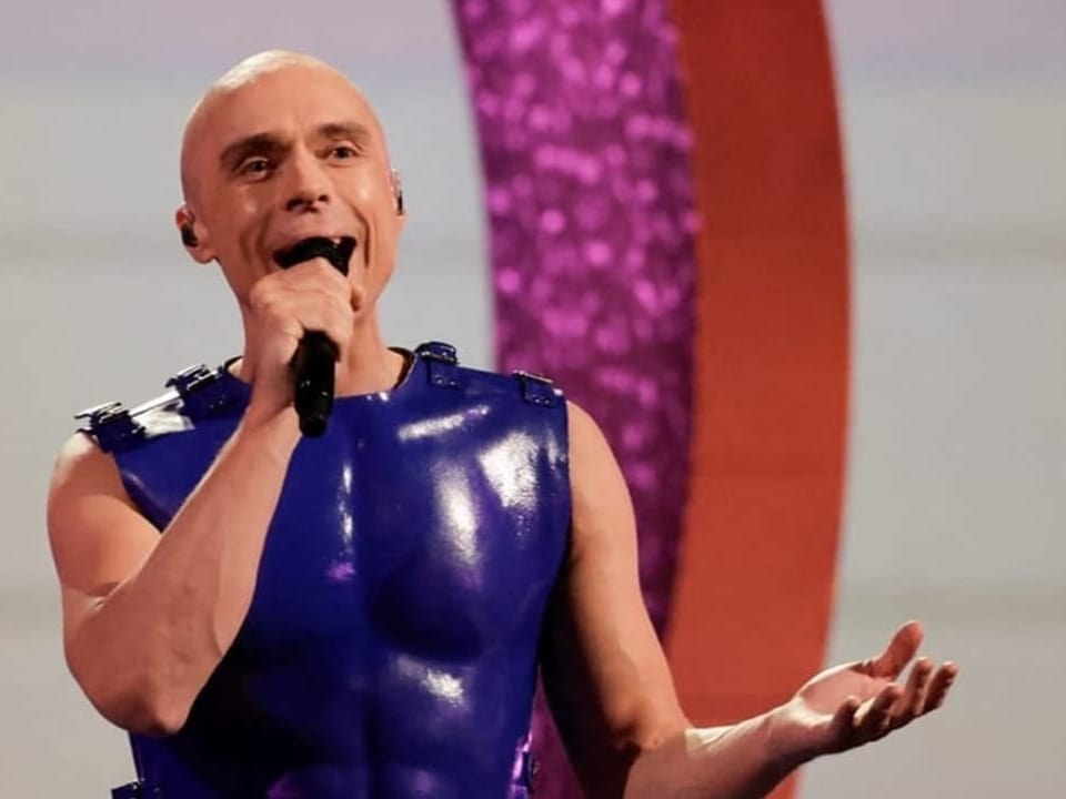 Glatzköpfiger Mann in blauem Oberleibkleid singt auf einer Bühne mit Mikrofon