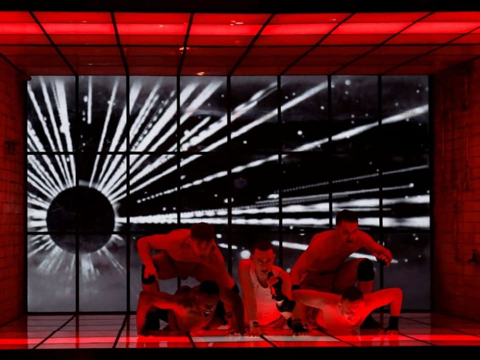 Dynamische Tanzszene in einem roten Raum mit futuristischem Videodisplay im Hintergrund.