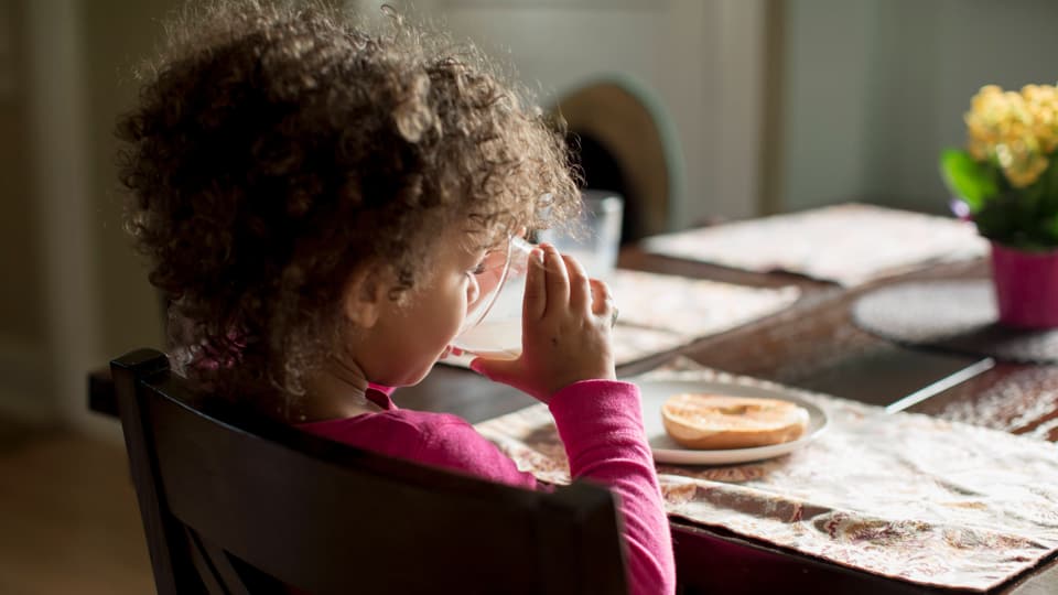 Little girl with curly hair drinks from a cup at the kitchen table.