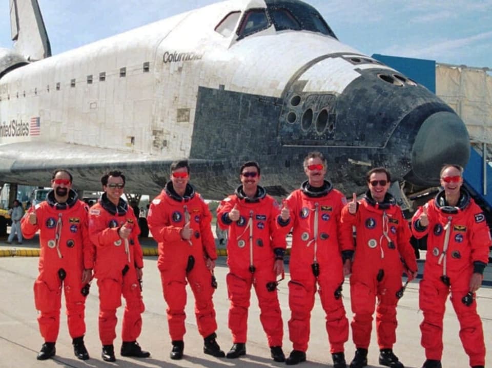 A group of seven astronauts in orange spacesuits in front of a space shuttle.