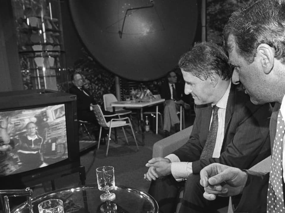 Two men look at a television showing a person in a spacesuit.
