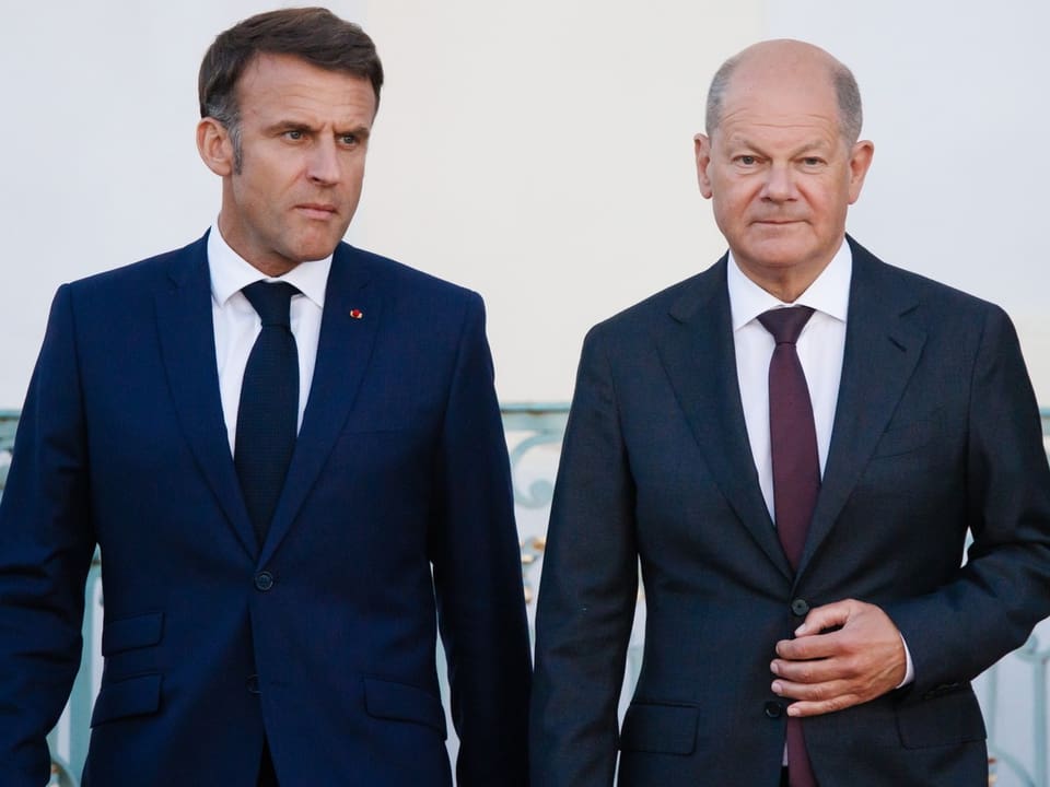 Left Macron in dark blue suit, critical look, right Scholz in dark grey suit, looking into camera