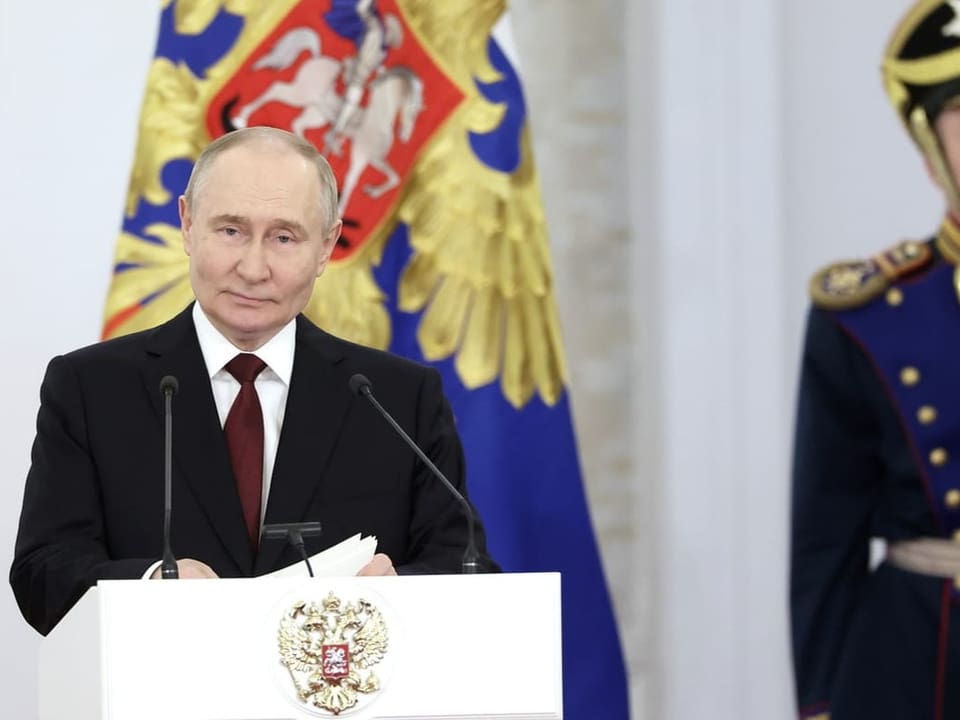 Putin at a white lectern, wearing a dark suit and dark red tie. Holding paper.