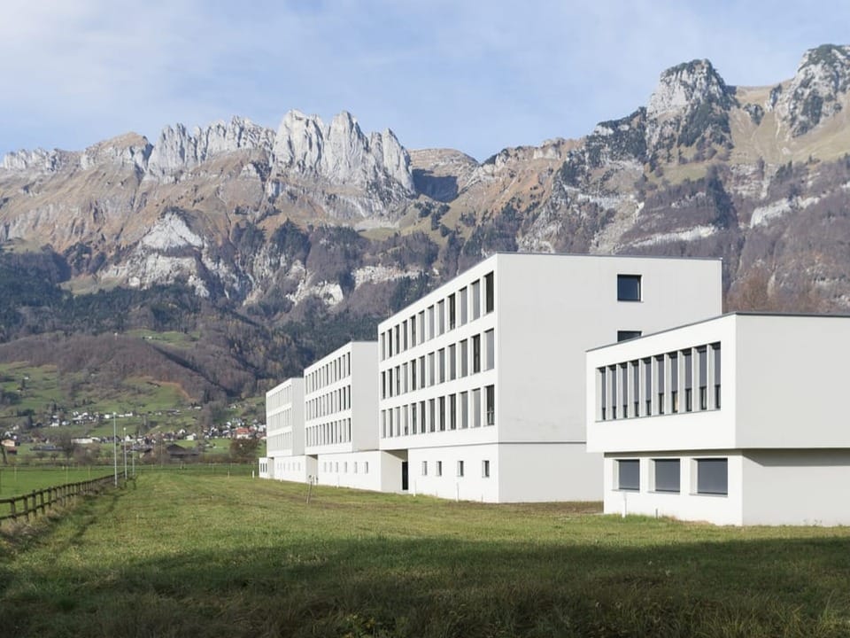 White concrete building, green meadows in the front, mountains in the background